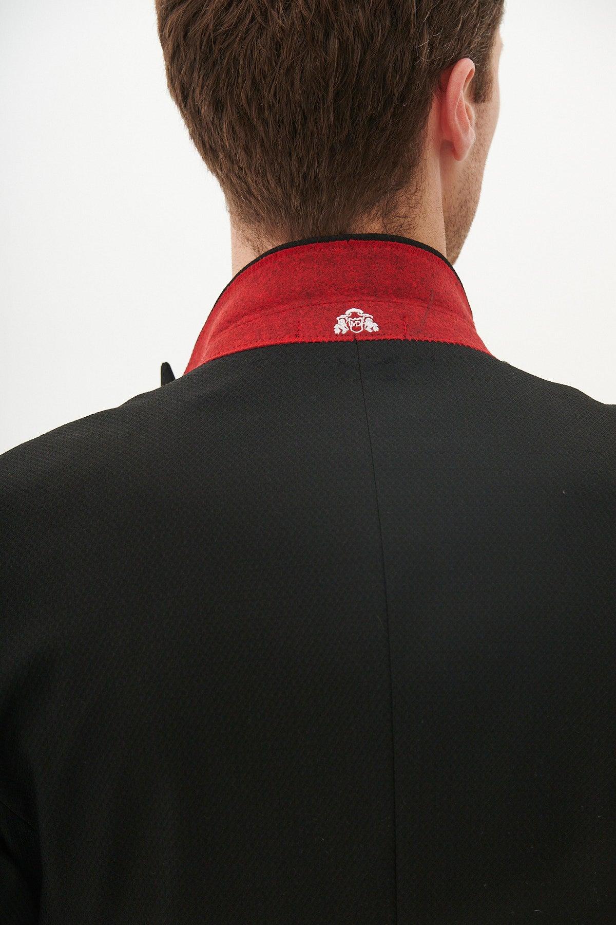 A man is seen from behind wearing the marcdarcy DALTON Black Tux Lapel Diamond Three Piece Suit, which has a red collar and a small white embroidered detail at the center. The background is plain and light-colored.