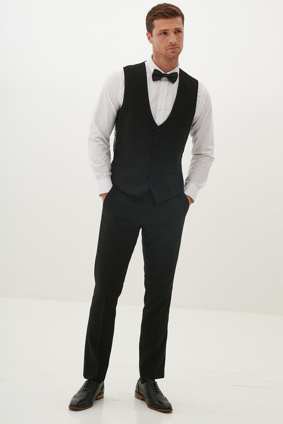 A man in a white shirt, black vest, bow tie, and pants with black dress shoes models the marcdarcy DALTON - Black Tux Lapel Diamond Three Piece Suit—a classic, elegant choice for formal occasions.