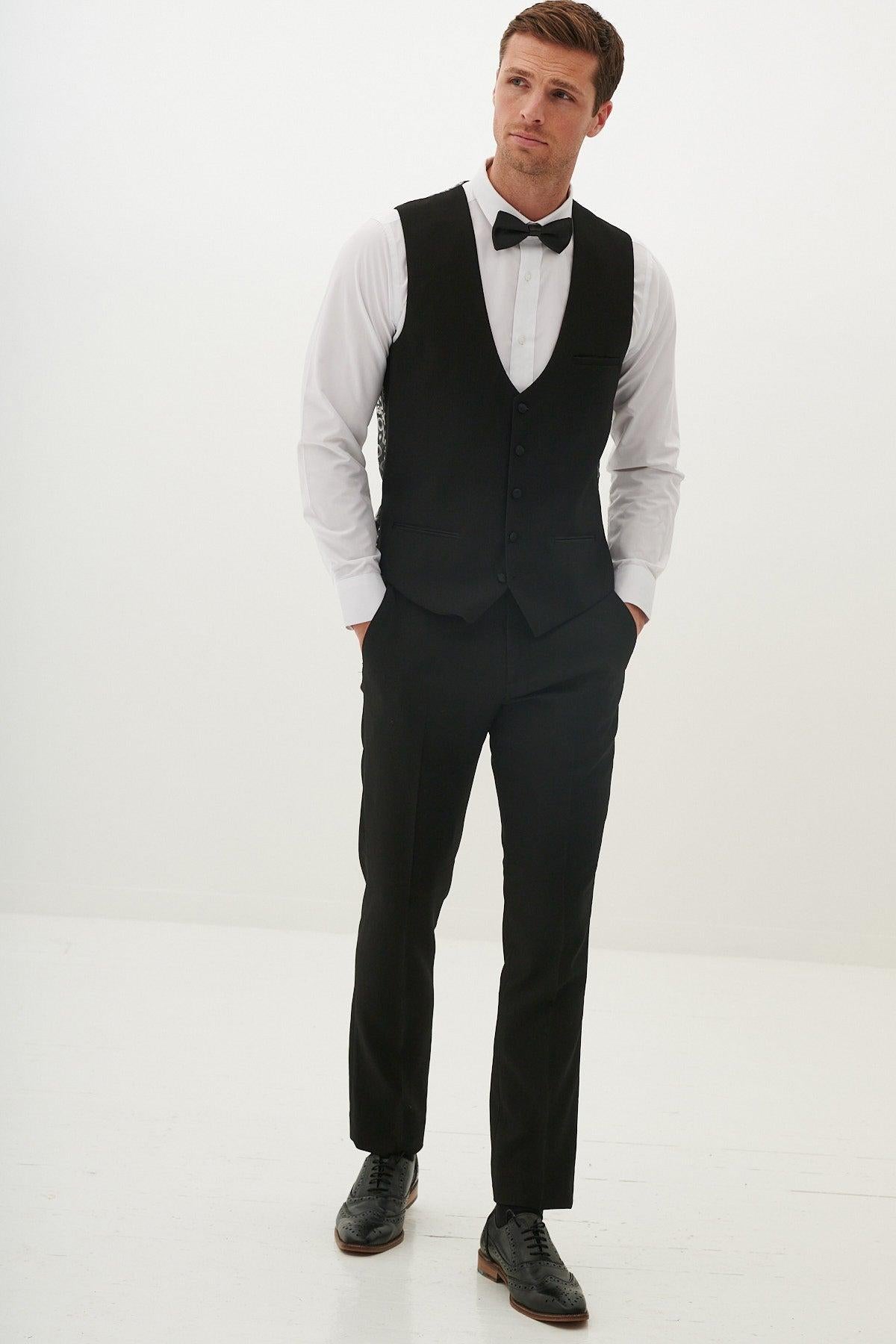 A man stands in a white room wearing the marcdarcy DALTON Black Tux Lapel Diamond Three Piece Suit with a vest, white shirt, black bow tie, and dress shoes. His hands are in his pants pockets as he looks slightly to the side.