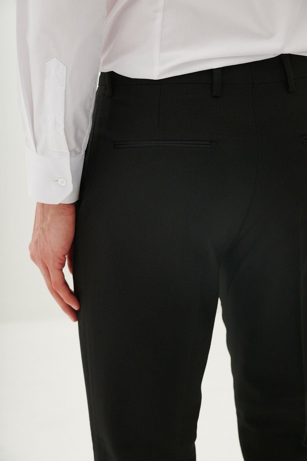 A person in the marcdarcy DALTON - Black Tux Lapel Diamond Three Piece Suit, featuring a white dress shirt and black dress pants, stands with their left hand by their side, shown from the back and waist down.