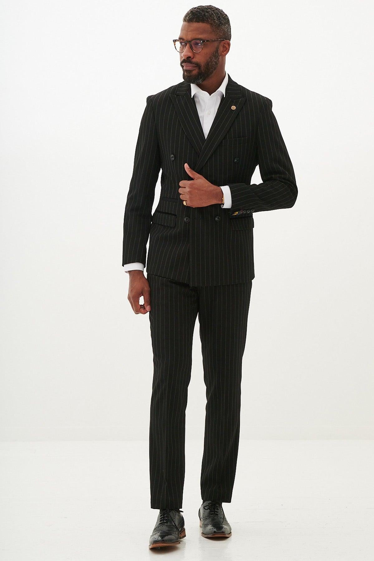 A man with short hair and a beard wears the Marc Darcy ROCCO Black Pinstripe Double Breasted Two Piece Suit with a white shirt, black shoes, and glasses, adjusting his jacket in a studio against a plain white background.