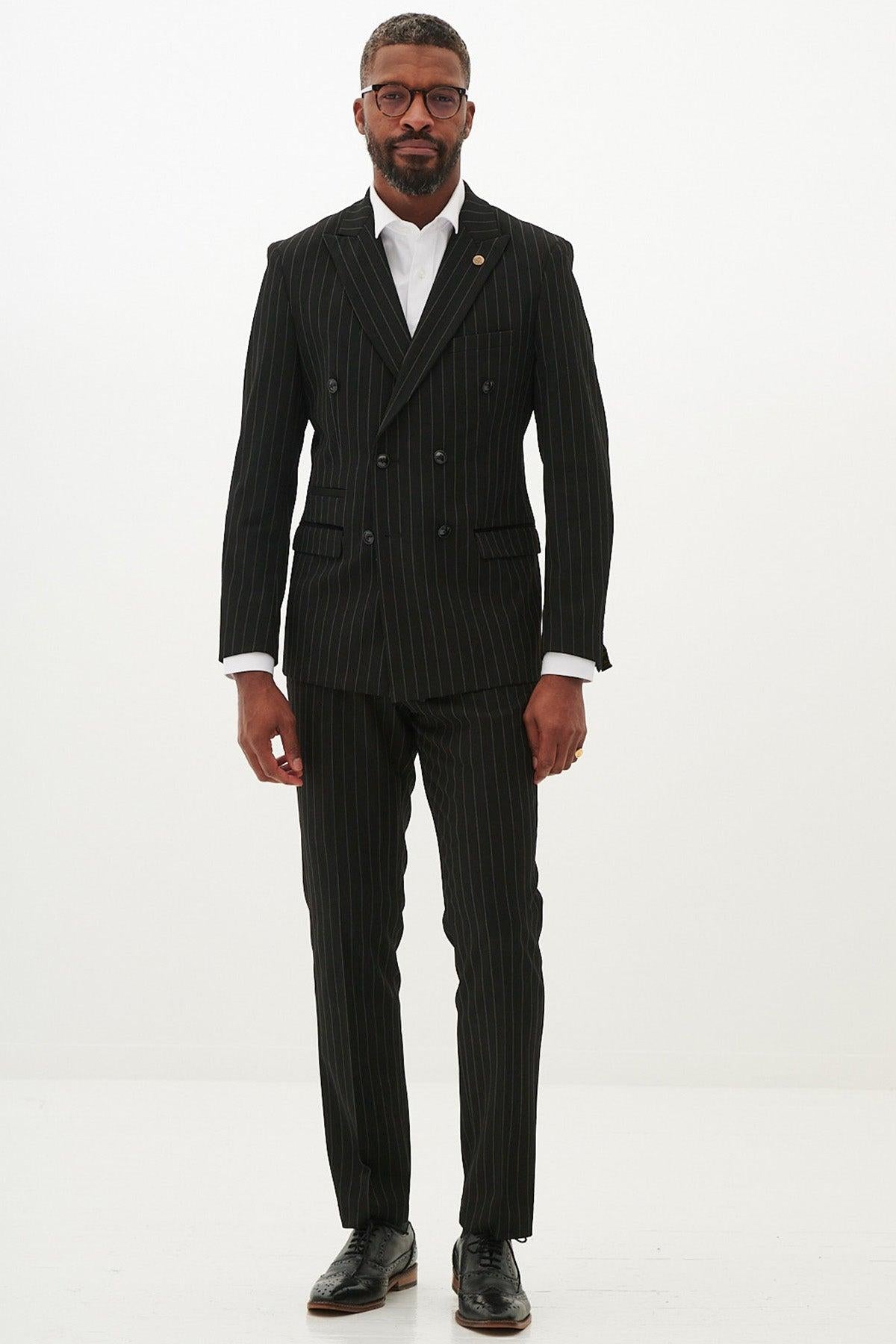 A man wearing the Marc Darcy ROCCO Black Pinstripe Double Breasted Two Piece Suit stands against a white background, paired with a white dress shirt and black shoes. He has short hair, glasses, a beard, and his hands rest by his sides.