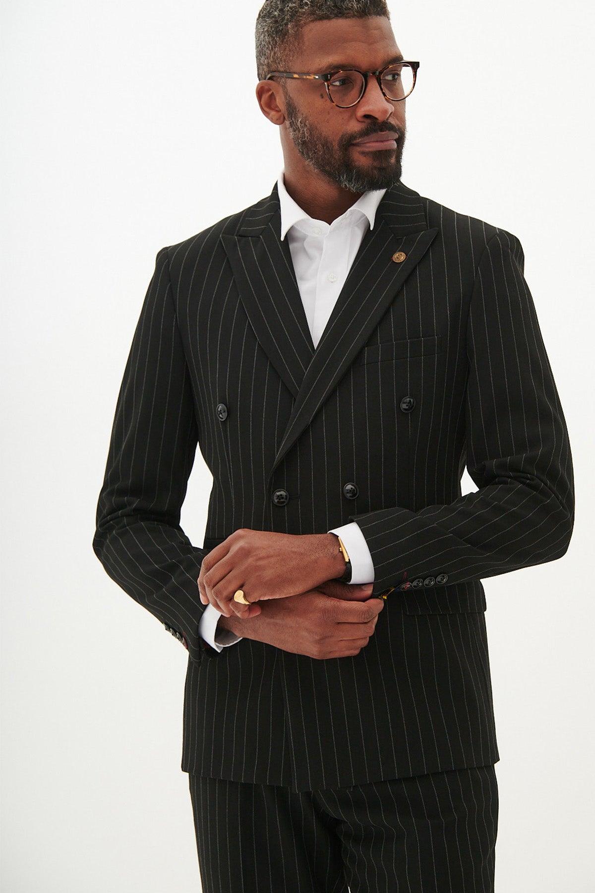 A man in glasses wears the Marc Darcy ROCCO Black Pinstripe Double Breasted Two Piece Suit with a white shirt, standing against a plain background while adjusting his sleeve and looking slightly to the side.