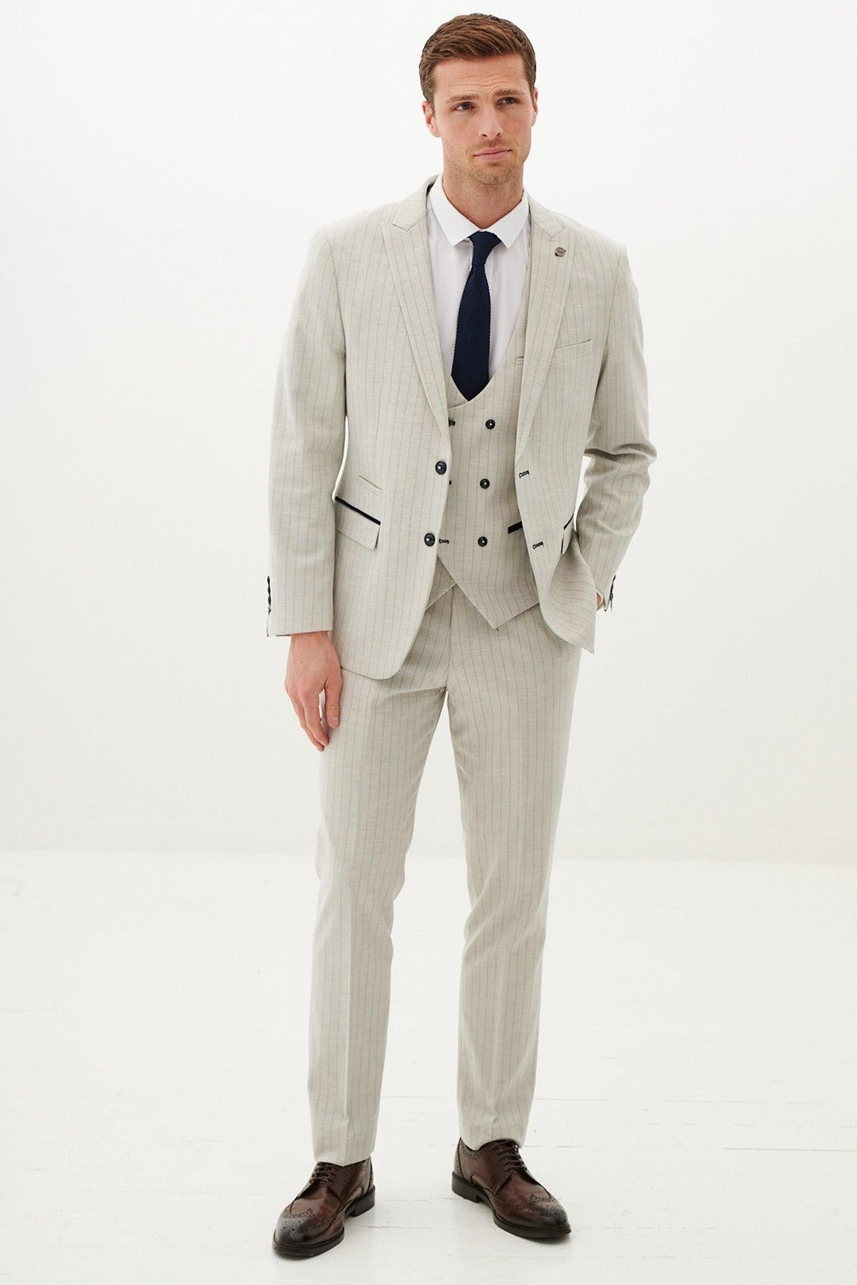 A man stands in a minimal white room wearing the Marc Darcy GRANT Stone Pinstripe Three Piece Suit with a white shirt, dark tie, and dark brown dress shoes, looking at the camera with a neutral expression.