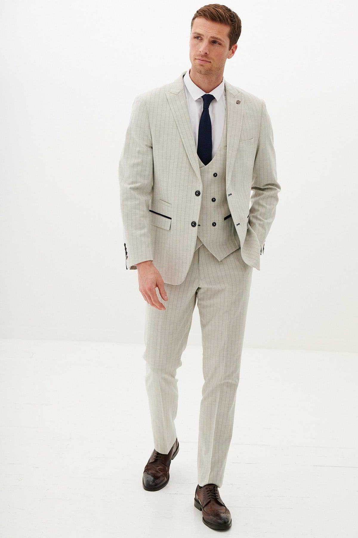 A man wears the Marc Darcy GRANT Stone Pinstripe Three Piece Suit with a white shirt, dark tie, and brown dress shoes. He stands against a plain white background, one hand in his pocket, gazing slightly to the side.