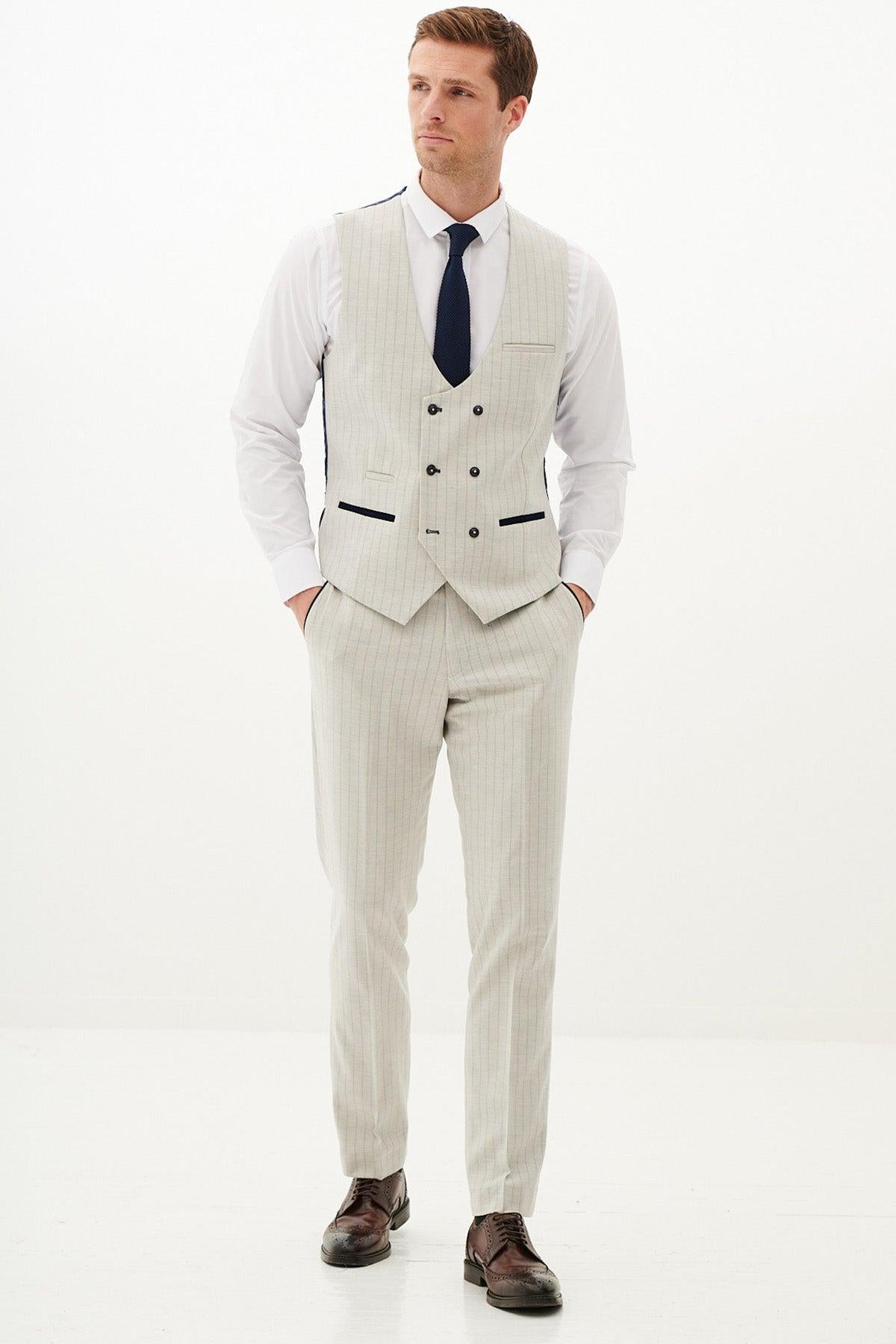 A man wears the Marc Darcy GRANT Stone Double Breasted Pinstripe Waistcoat with a white shirt, navy tie, and brown dress shoes, standing hands-in-pockets against a plain white background.