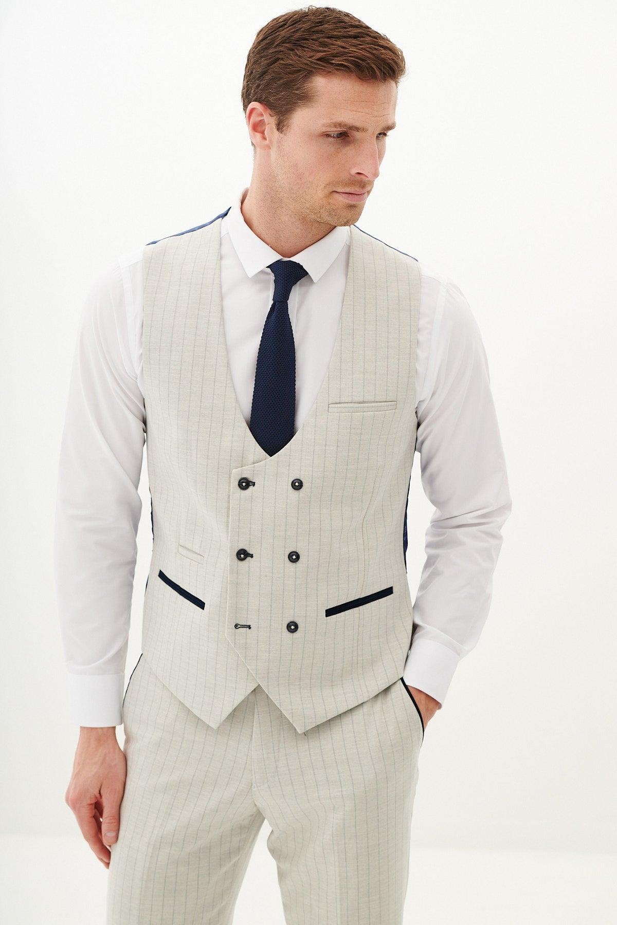 A man models the Marc Darcy GRANT Stone Pinstripe Three Piece Suit, pairing it with a white dress shirt and dark tie, standing with one hand in his pocket against a plain white background.