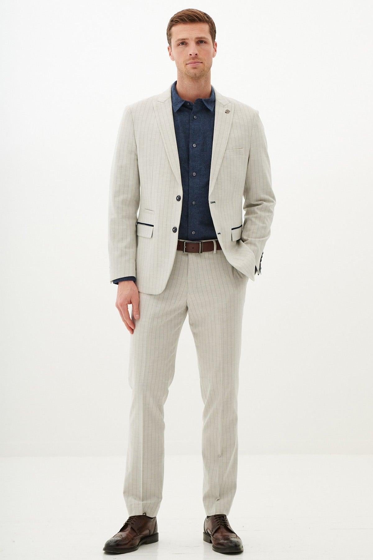 A man faces forward with a neutral expression and his left hand in his pocket, wearing the Marc Darcy GRANT - Stone Pinstripe Two Piece Suit, paired with a dark blue shirt, brown belt, and brown dress shoes against a plain white background.