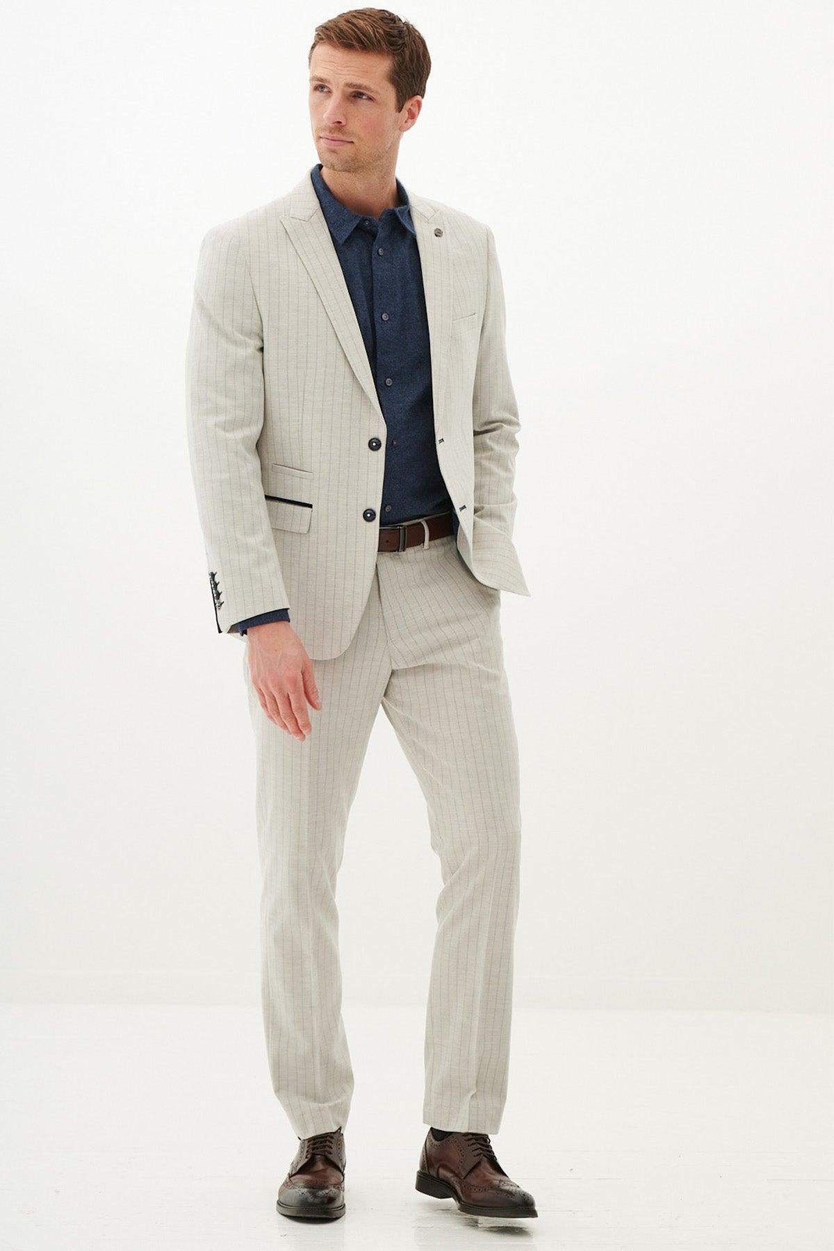A man stands against a plain white background, wearing the Marc Darcy GRANT Stone Pinstripe Blazer with navy accents, paired with a brown belt and brown dress shoes. His hands are relaxed, and he gazes slightly to the side.