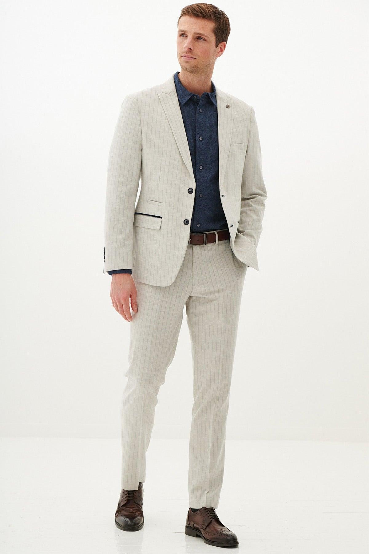 A man stands against a plain white background wearing the Marc Darcy GRANT - Stone Pinstripe Blazer with a dark blue shirt, brown belt, and brown dress shoes. With one hand in his pocket, he gazes slightly to the side.