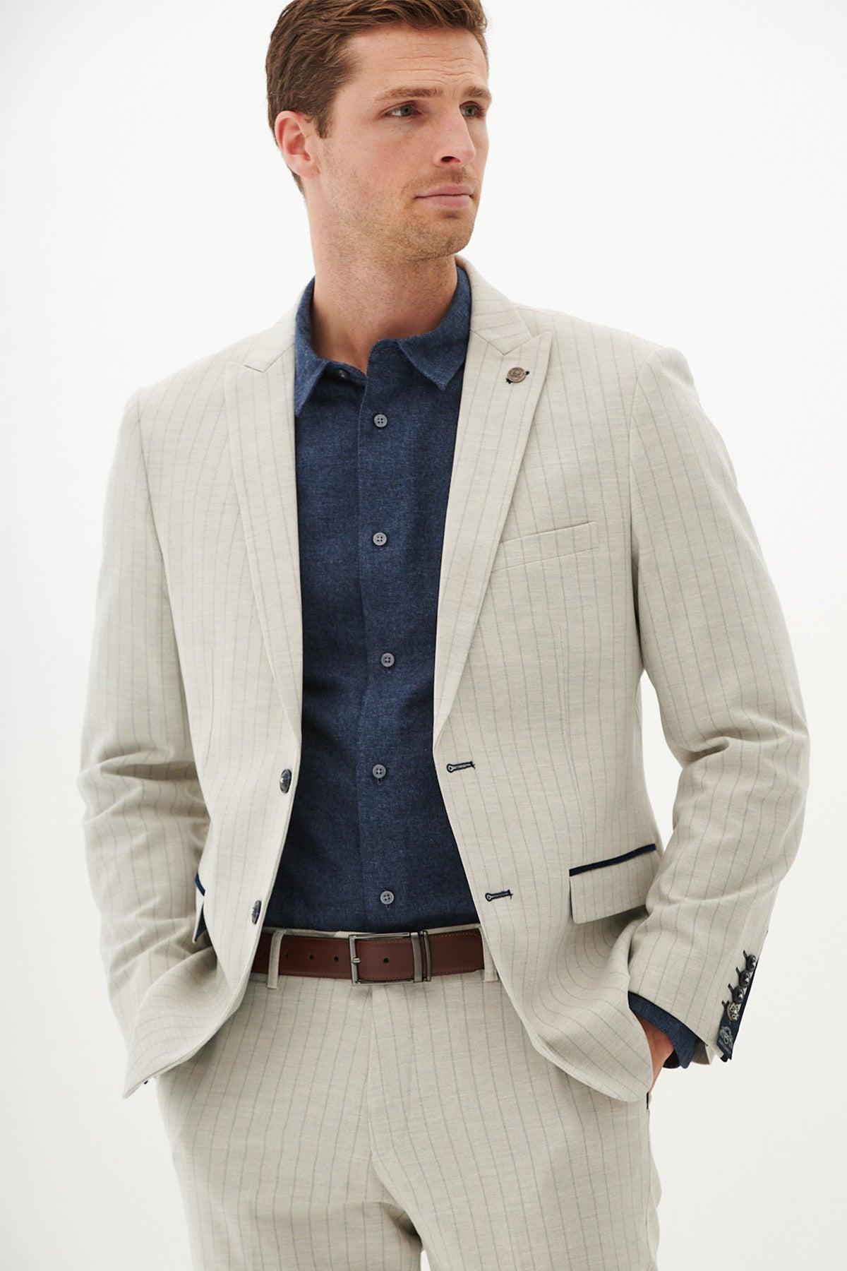 A man wears the GRANT - Stone Pinstripe Blazer by Marc Darcy with a navy blue button-down shirt and a brown belt.