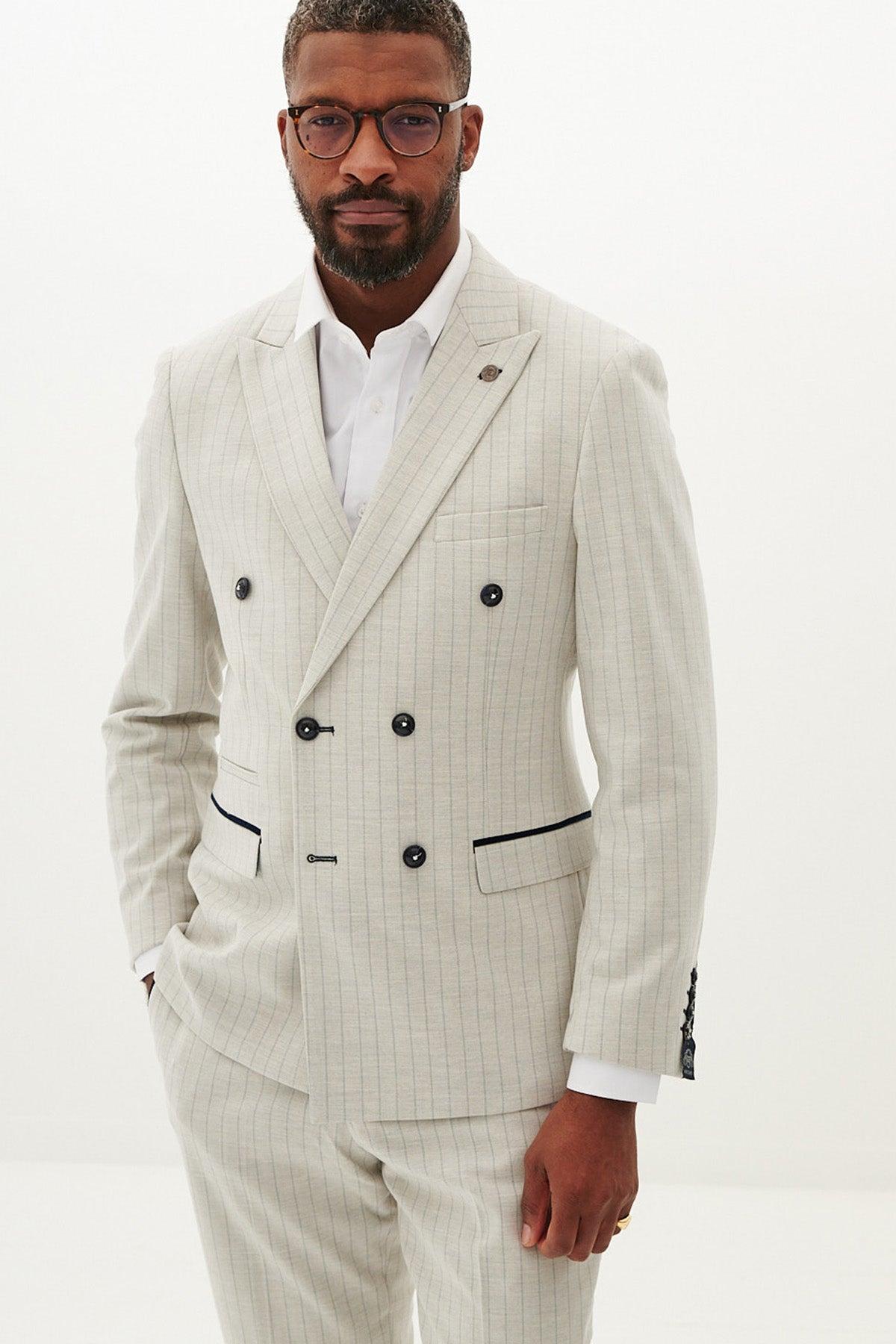 A man wearing glasses models the Marc Darcy GRANT - Stone Double Breasted Pinstripe Blazer, featuring black buttons and trim, standing against a plain white background with one hand in his pocket.