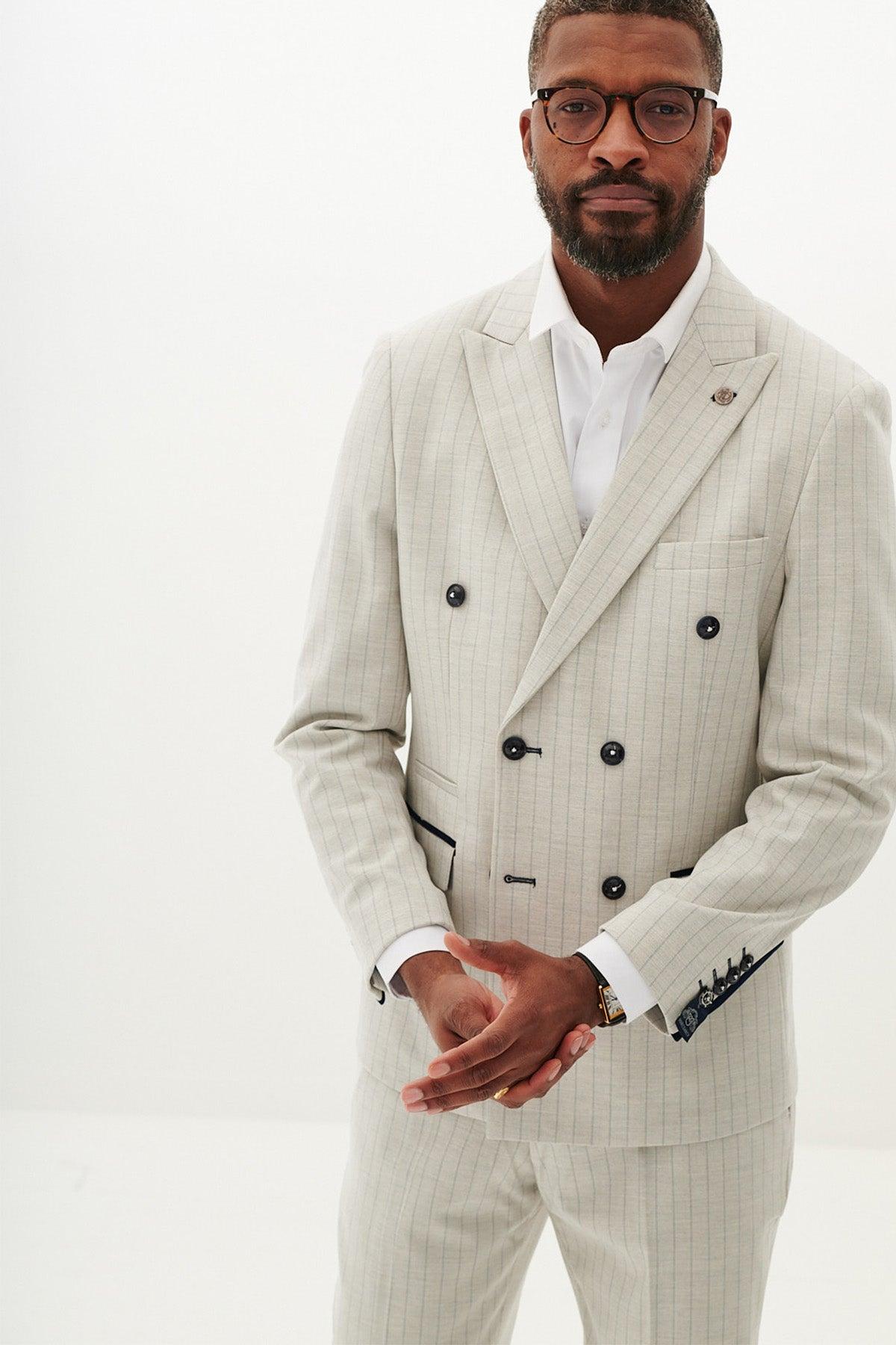 A man in the Marc Darcy GRANT Stone Pinstripe Double Breasted Suit with a white shirt stands against a plain white background, wearing glasses and looking at the camera with his hands clasped.