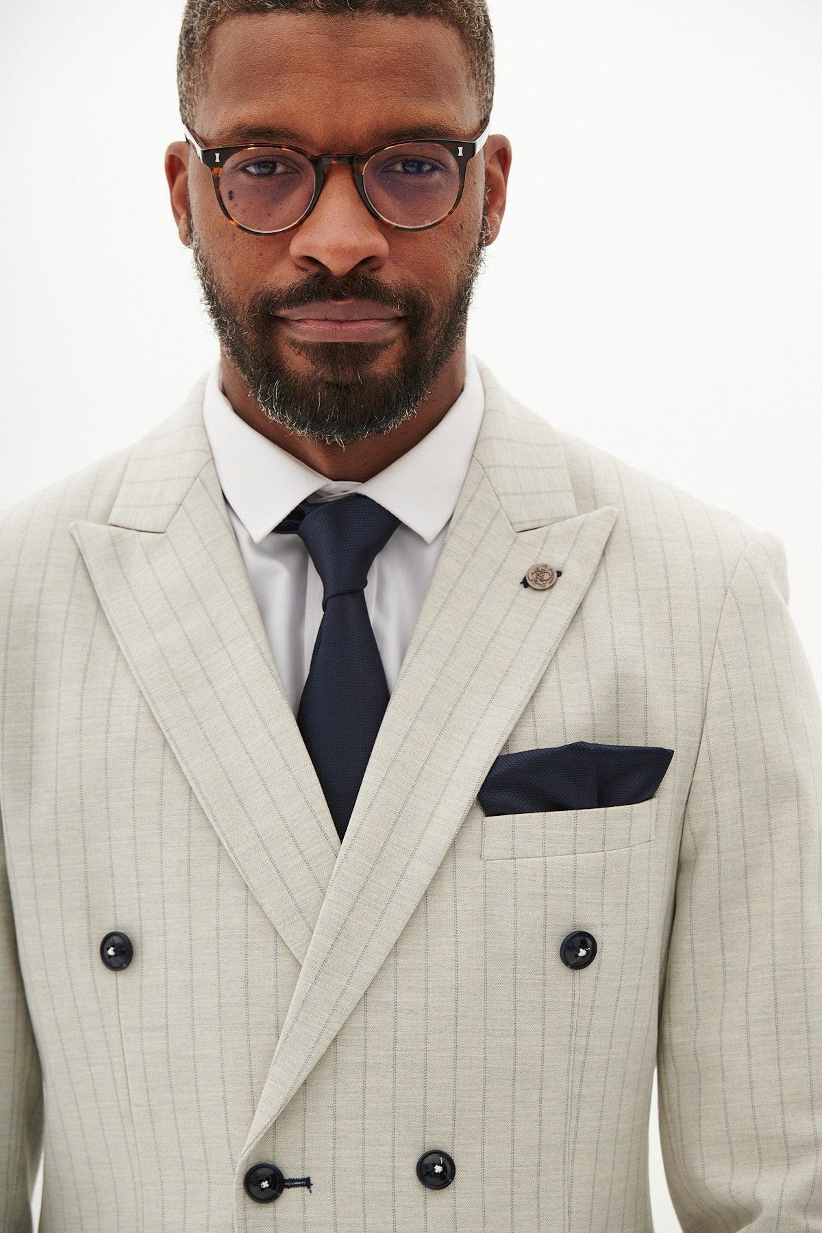 A man in the Marc Darcy GRANT Stone Double Breasted Pinstripe Blazer, paired with a white shirt, navy tie, and matching pocket square, stands against a plain white background.