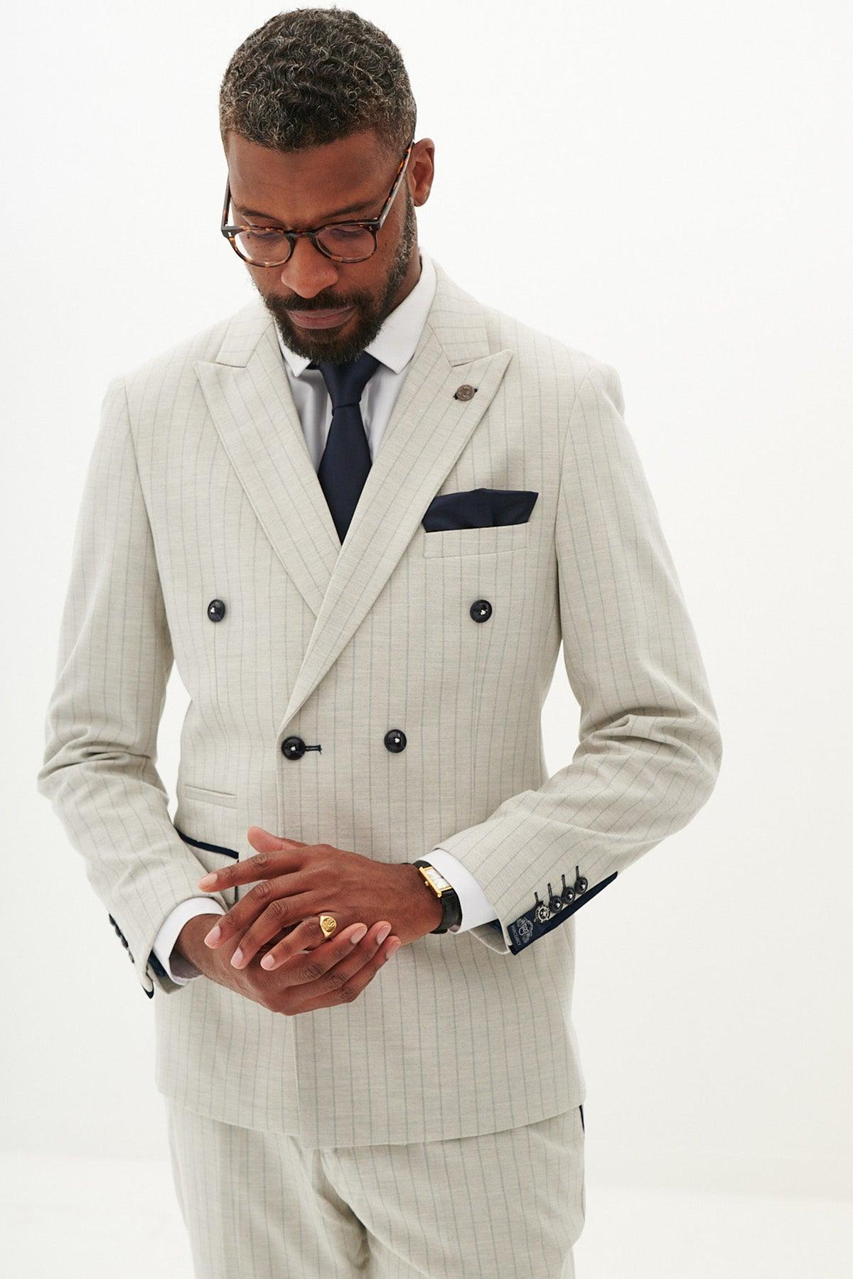 A man wearing the Marc Darcy GRANT - Stone Double Breasted Pinstripe Blazer, white shirt, navy tie, glasses, ring, and a navy pocket square adjusts his cuff against a plain white background.