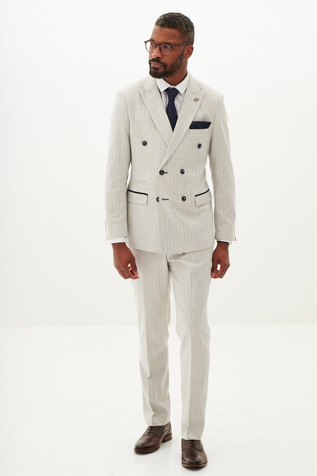 A man wearing the Marc Darcy GRANT - Stone Pinstripe Double Breasted Suit with black buttons, a white shirt, dark tie, brown shoes, and glasses stands against a plain white background.