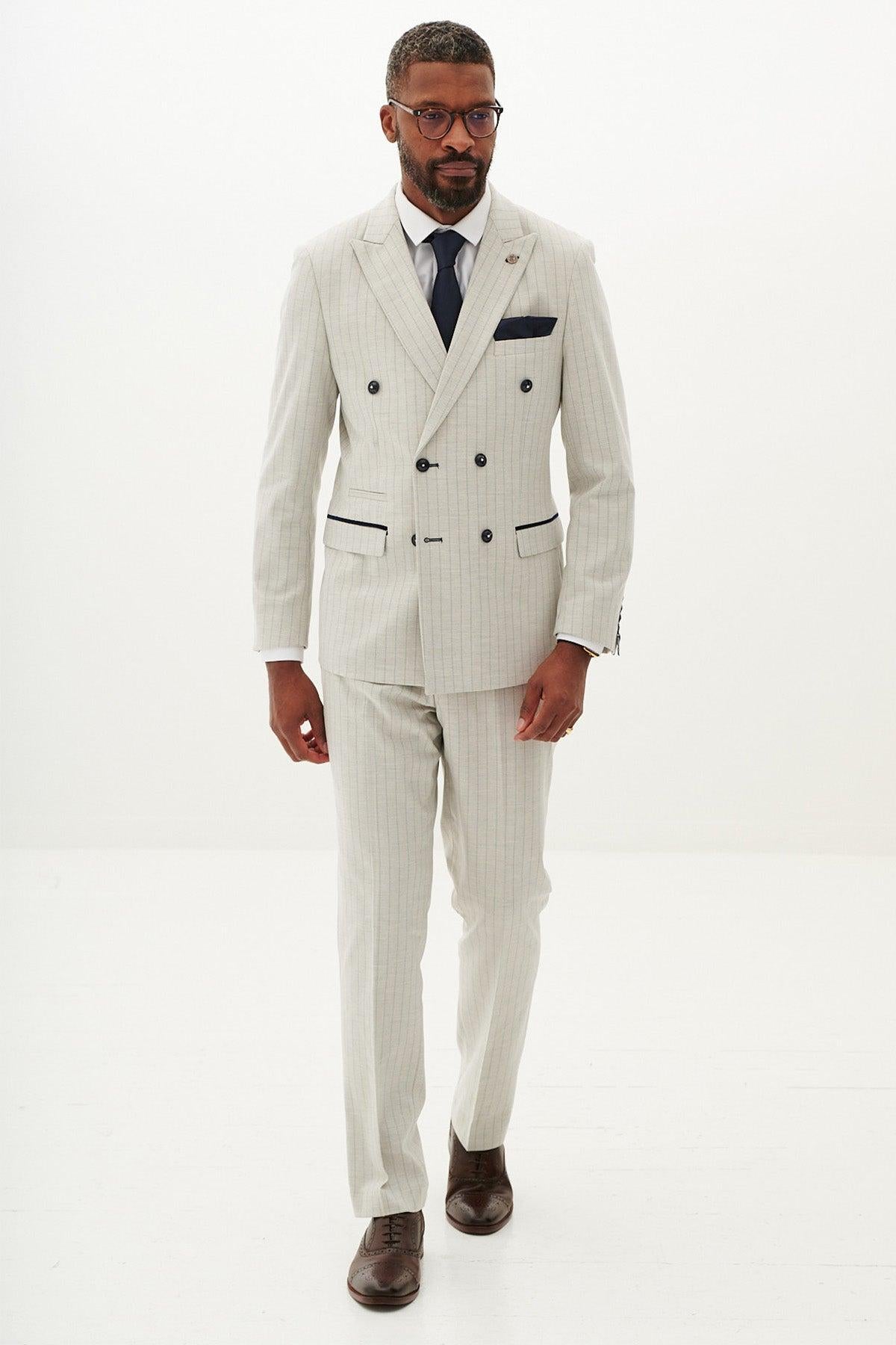 A man wears the Marc Darcy GRANT Stone Pinstripe Double Breasted Suit—a slim fit gray pinstripe three-piece with black buttons, white shirt, dark tie, black pocket square, and brown shoes—against a plain white background.