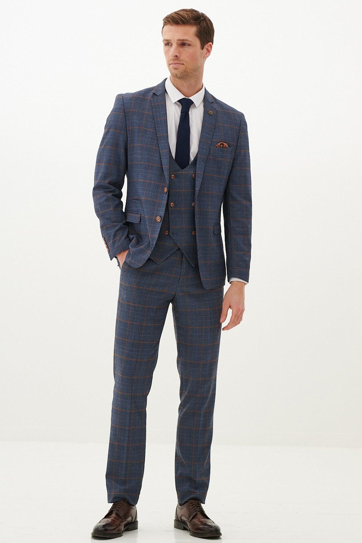 A man stands against a plain background wearing the marcdarcy JENSON sky blue check suit with a double-breasted waistcoat, paired with a white shirt, navy tie, brown dress shoes, and a patterned pocket square. He looks to the side with one hand by his side.