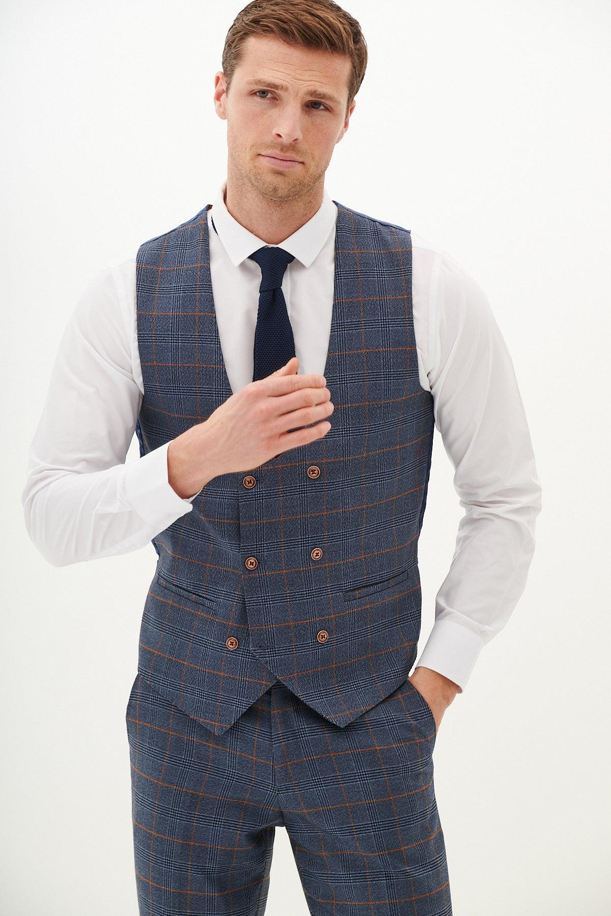 A man dressed in the marcdarcy JENSON Sky Blue Check Suit with Double Breasted Waistcoat, white shirt, and navy tie stands against a plain white background, one hand on his chest and the other in his pocket.