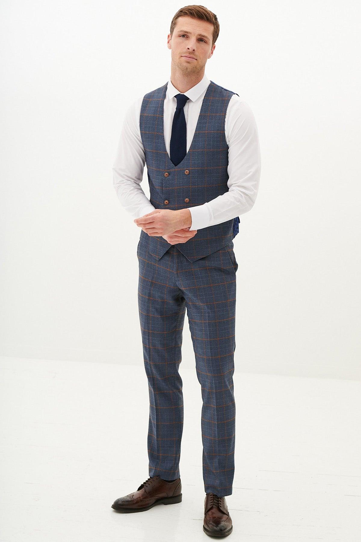 A man with short hair and a neutral expression stands against a white background, wearing the marcdarcy JENSON Sky Blue Check Double Breasted Waistcoat over a white shirt, navy tie, and brown shoes; his hands are clasped in front.