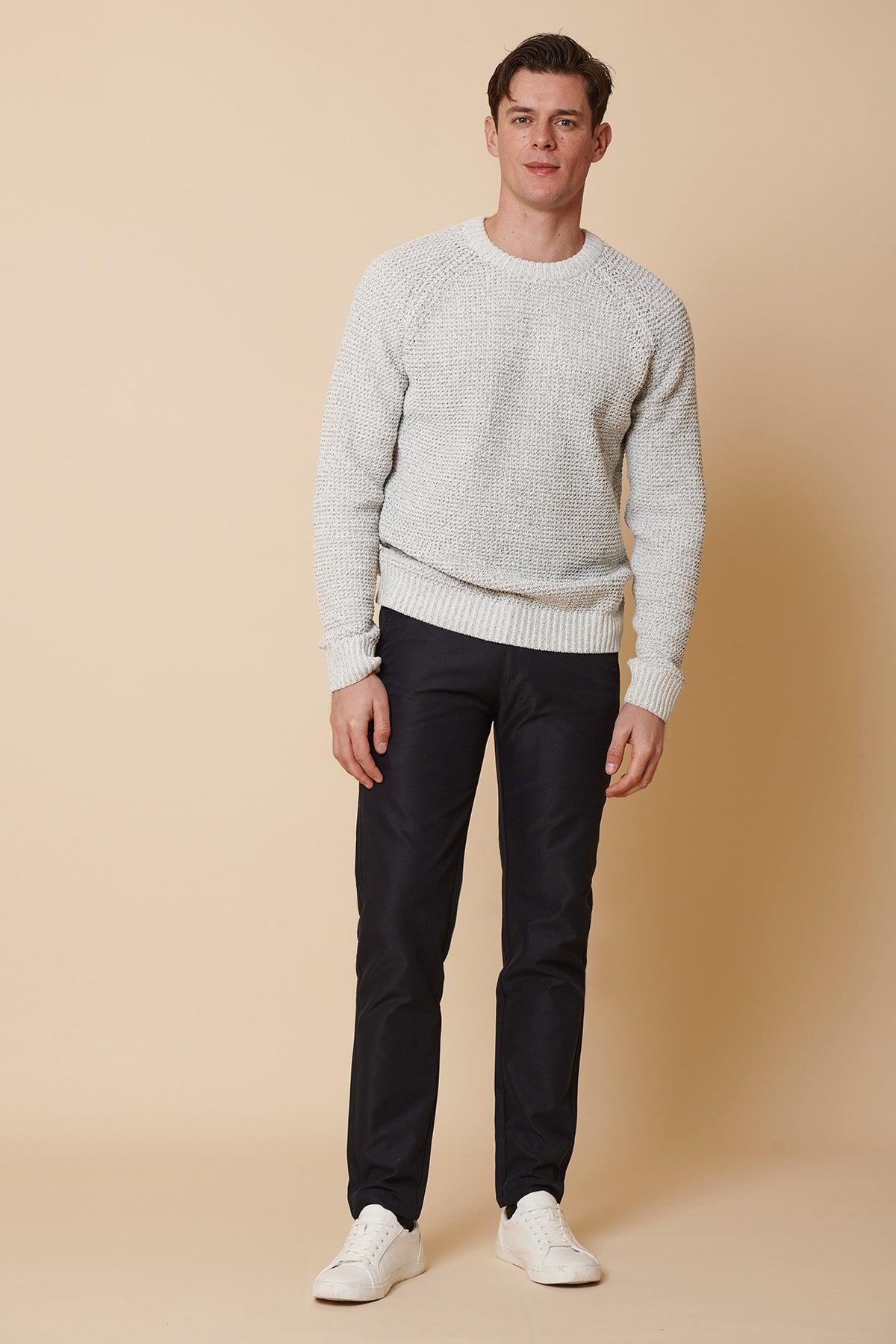 A man stands against a beige background wearing marcdarcys ENZO Navy Chinos, a light gray knit sweater, and white sneakers, looking at the camera with a relaxed posture.