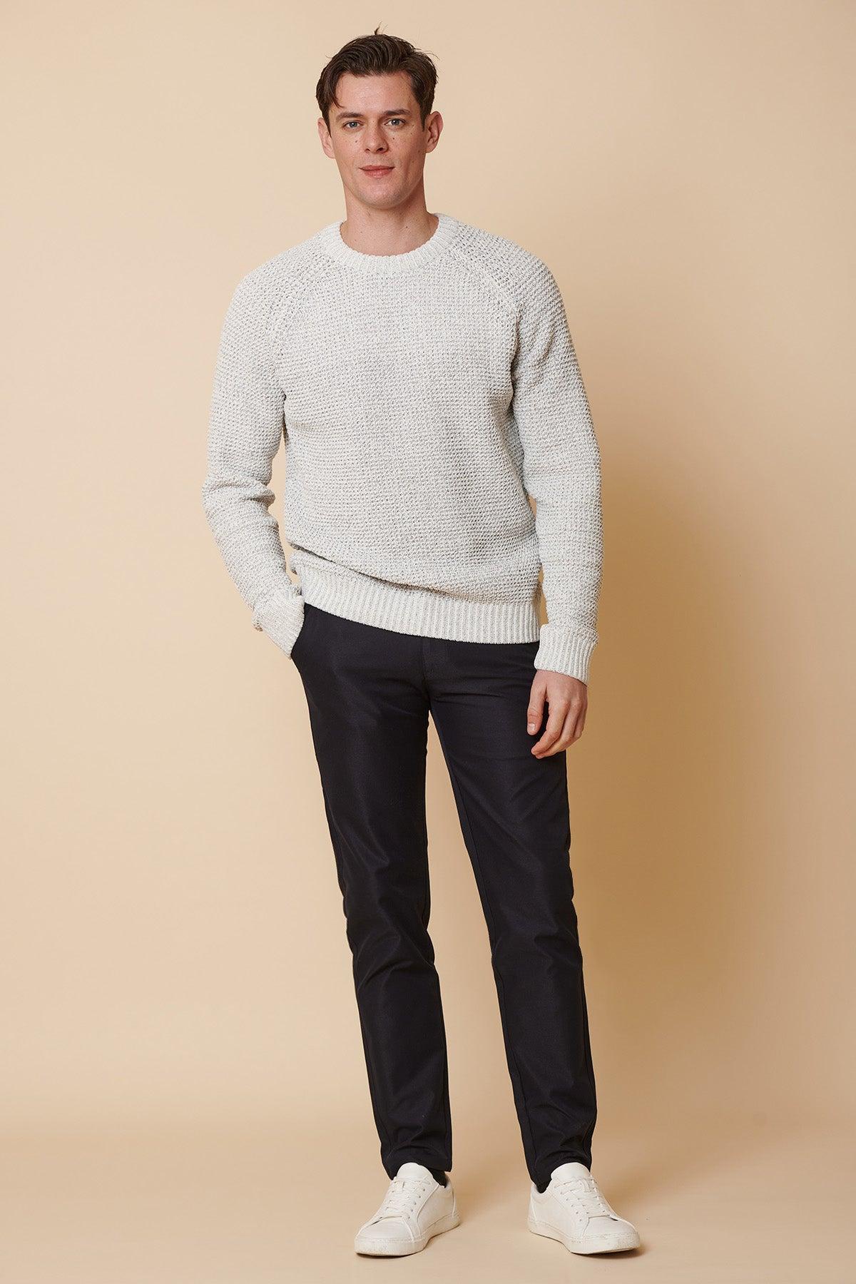 A man stands with hands in pockets against a beige background, wearing marcdarcy ENZO Navy Chinos, a light grey textured sweater, and white sneakers.