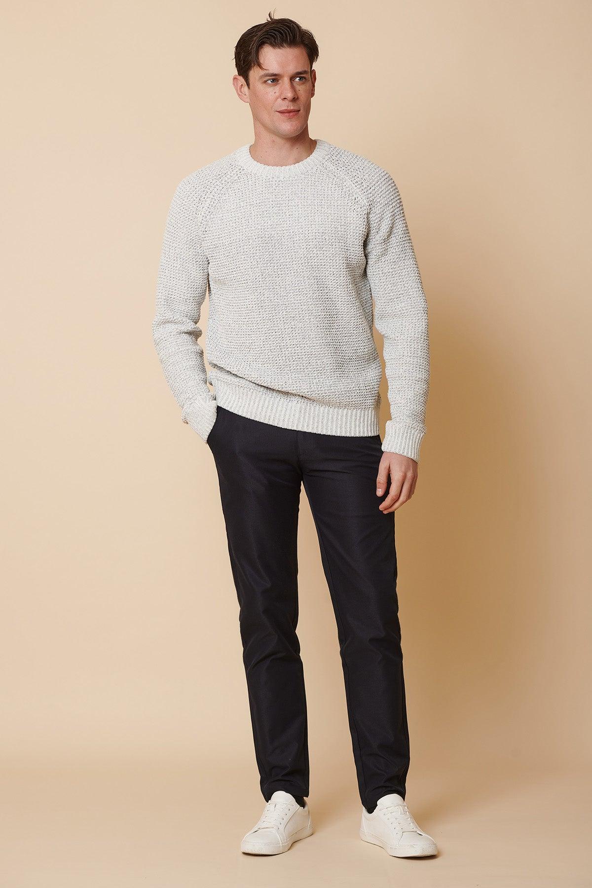 A man stands against a beige background wearing marcdarcy ENZO Navy Chinos, a light gray textured sweater, and white sneakers. He has short brown hair and stands with one hand in his pocket, looking slightly to the side.