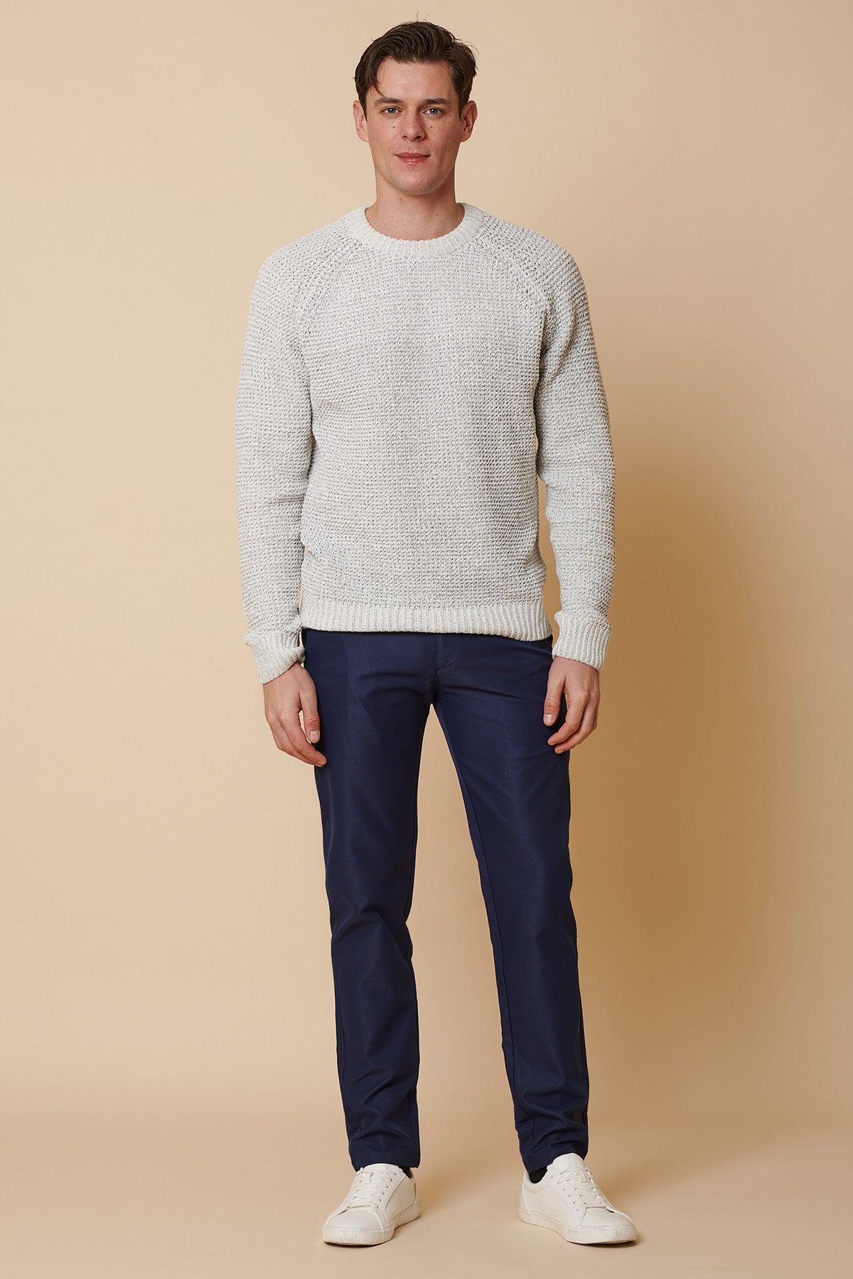 A man stands against a beige background wearing marcdarcy’s ENZO Royal Blue Chinos, a light gray textured sweater, and white sneakers. He faces forward with a neutral expression, arms relaxed at his sides.