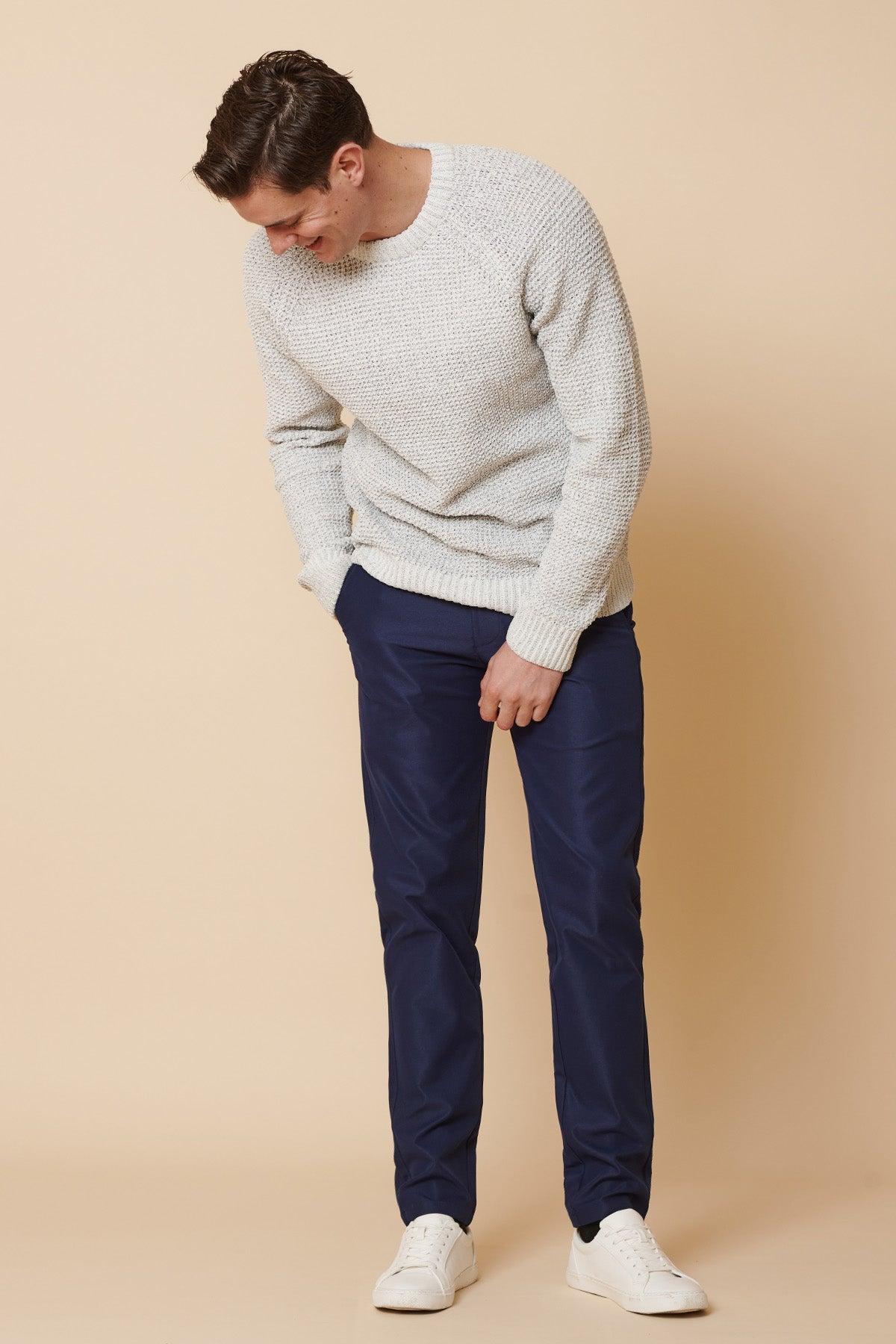 A man wearing a light gray knit sweater, marcdarcy ENZO Royal Blue Chinos, and white sneakers stands against a beige background, looking down with one hand in his pocket and the other adjusting his pants.