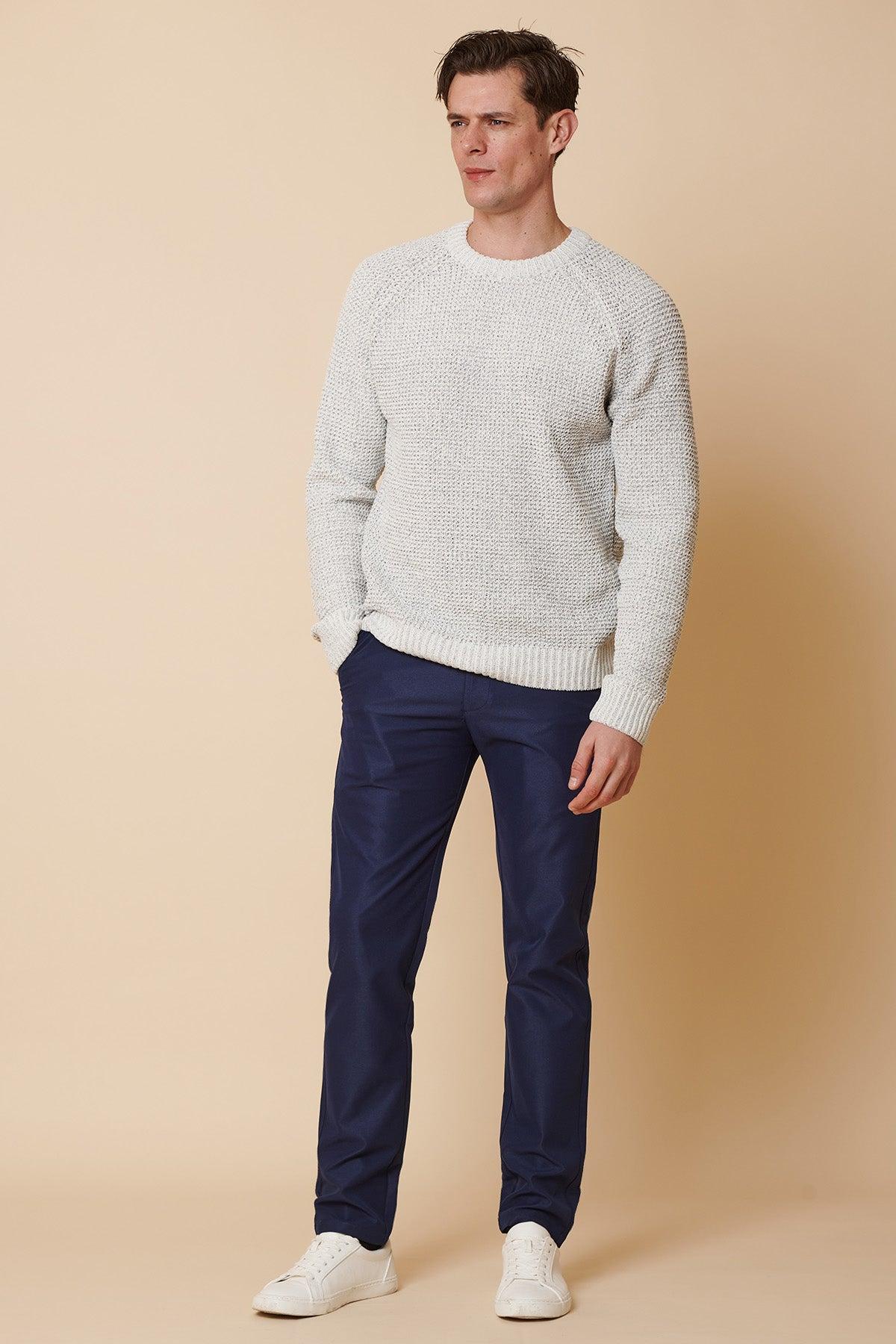 A man stands against a beige background wearing a light gray knit sweater, marcdarcy ENZO Royal Blue Chinos, and white sneakers. His left hand is in his pocket as he looks slightly to the right.