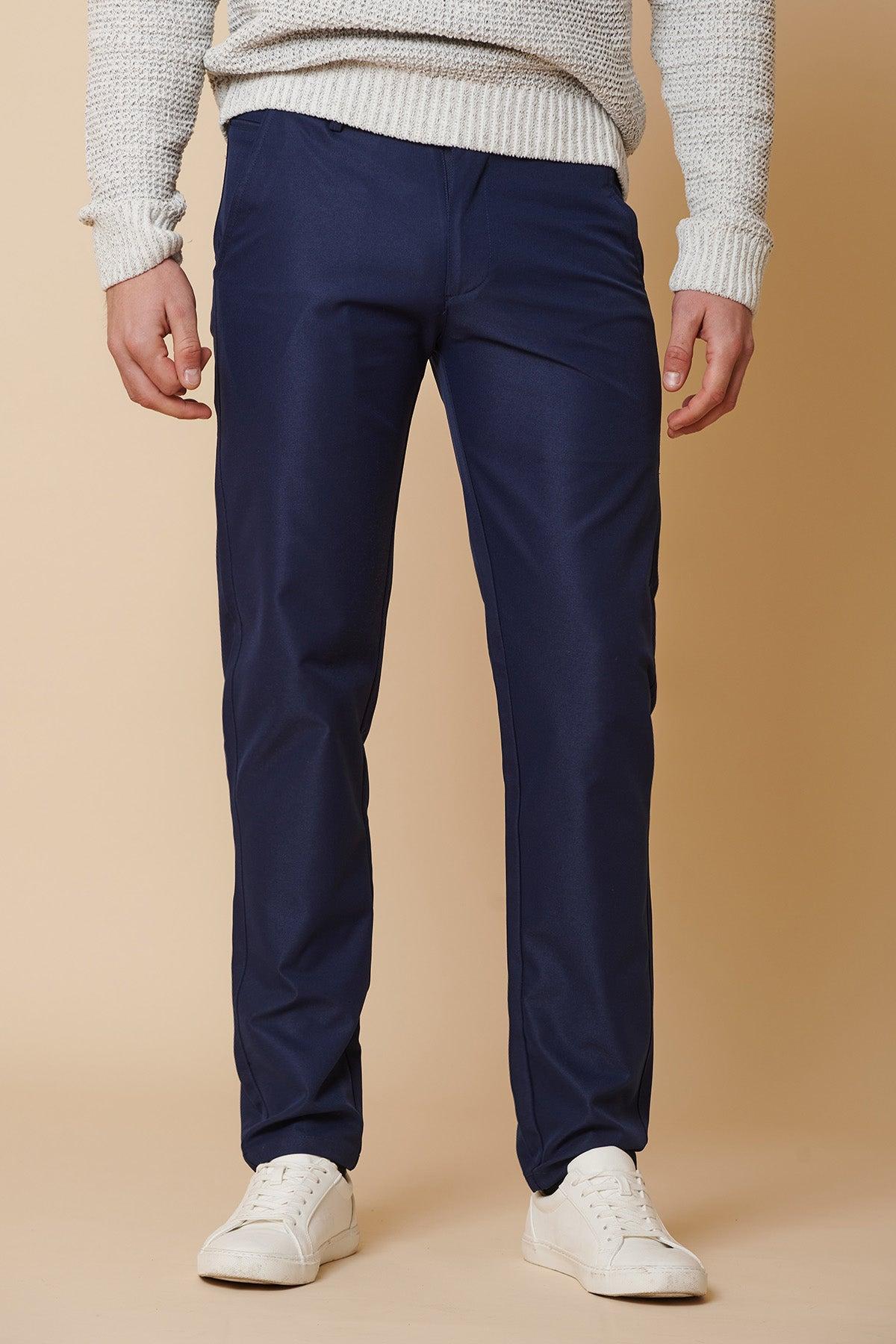 Against a beige background, only the lower half is shown: a person wears marcdarcy's ENZO Royal Blue Chinos paired with a light textured sweater and white sneakers.