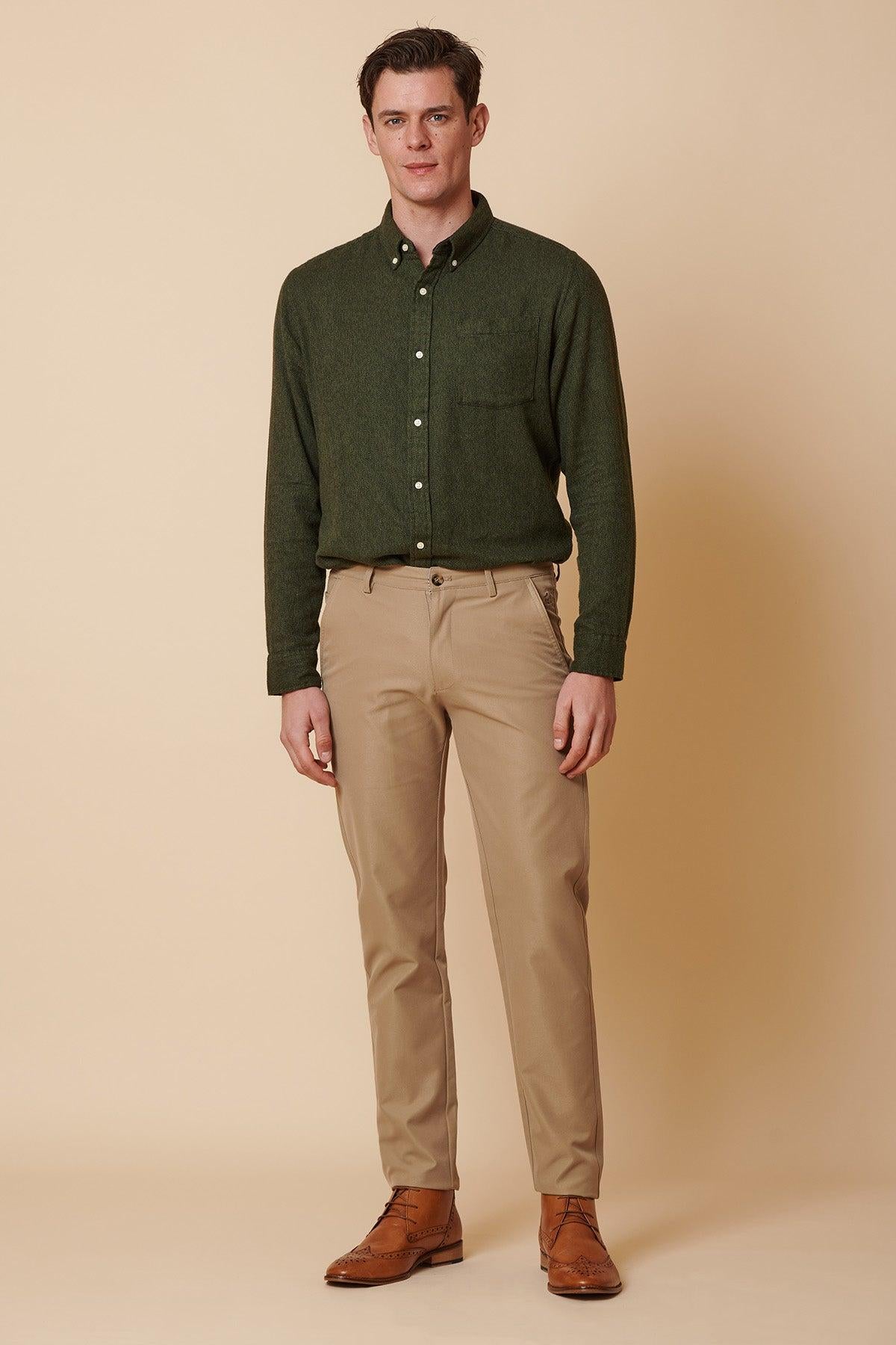 A man stands against a beige background wearing marcdarcy's ENZO - Sand Chinos, a dark green button-up shirt, and brown dress shoes. He faces the camera with a neutral expression and his hands by his sides.