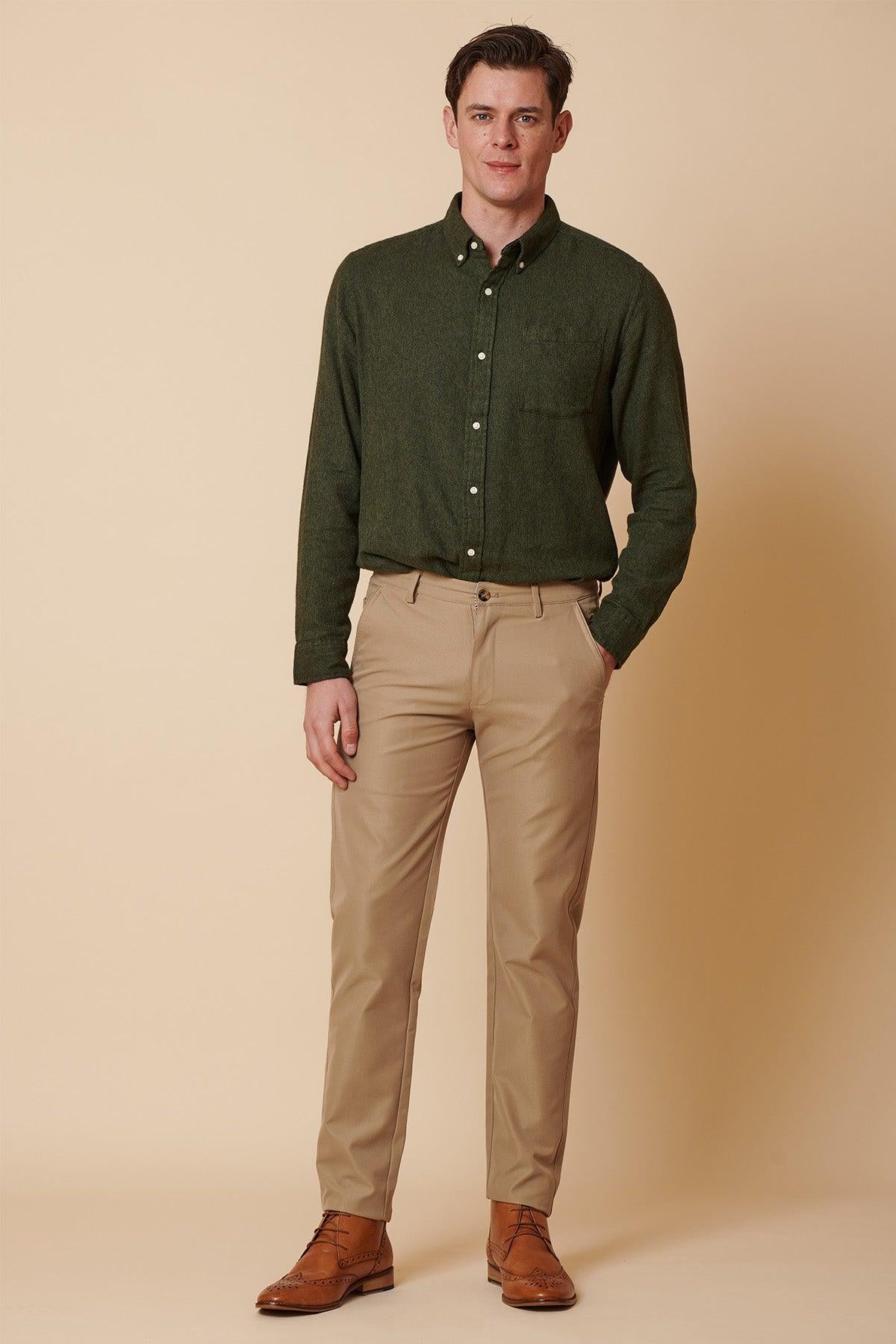 A man stands against a beige background, wearing marcdarcy ENZO - Sand Chinos, a dark green button-up shirt, and brown dress shoes. One hand is in his pocket as he looks at the camera with a neutral expression.