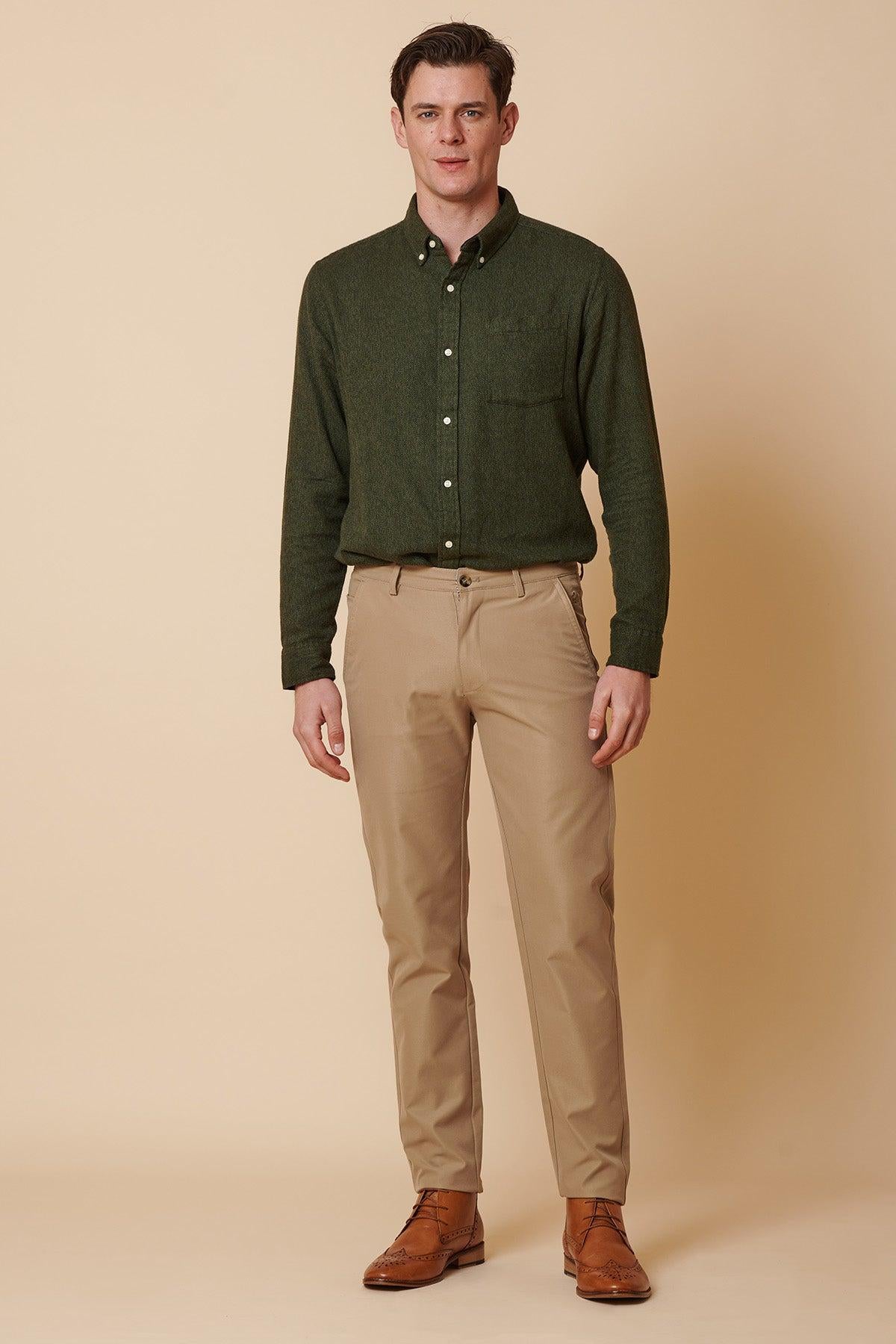 A man stands against a beige background, wearing marcdarcy's ENZO - Sand Chinos paired with a dark green button-up shirt and brown dress shoes. He faces forward with a neutral expression and relaxed posture.
