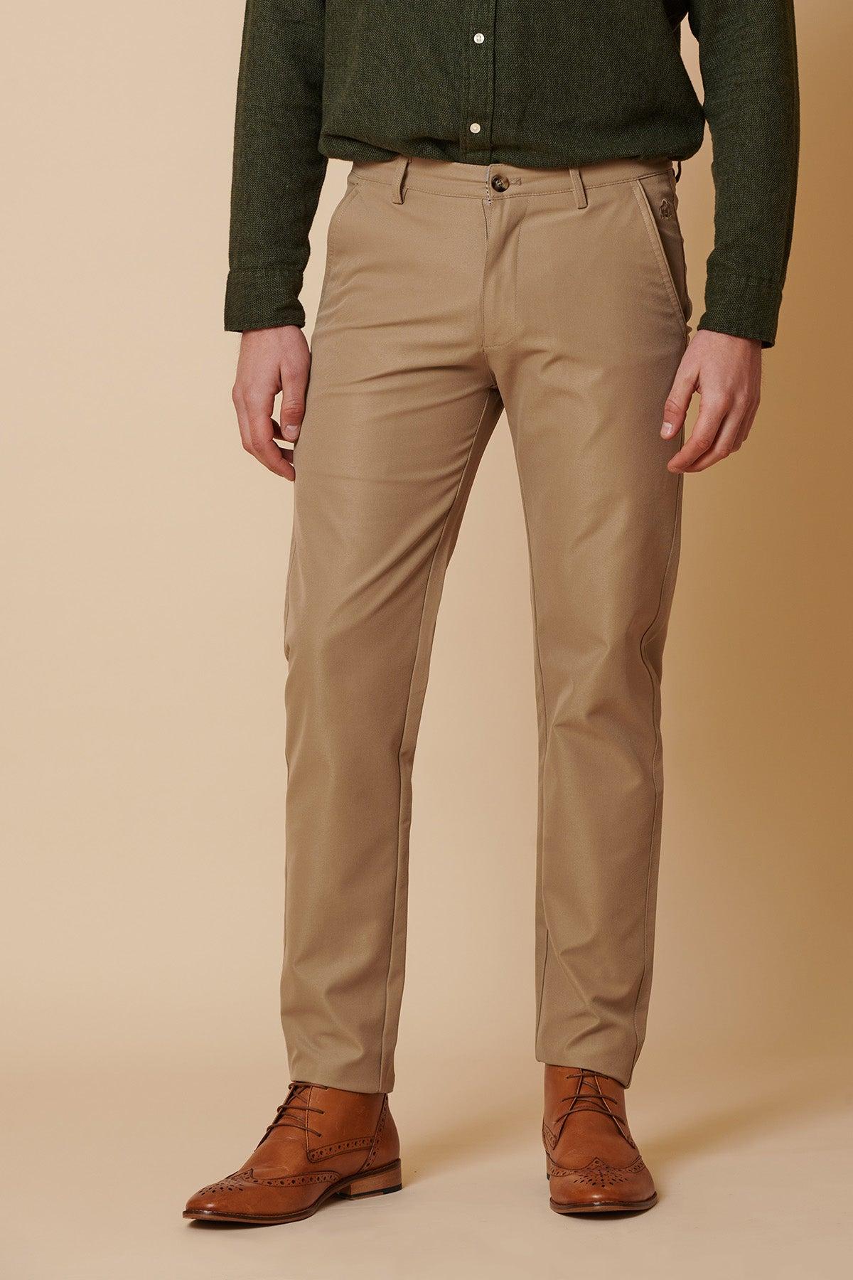 A person wears marcdarcy ENZO - Sand Chinos, a dark green shirt, and brown leather dress shoes against a beige background. Their head is not visible in the image.