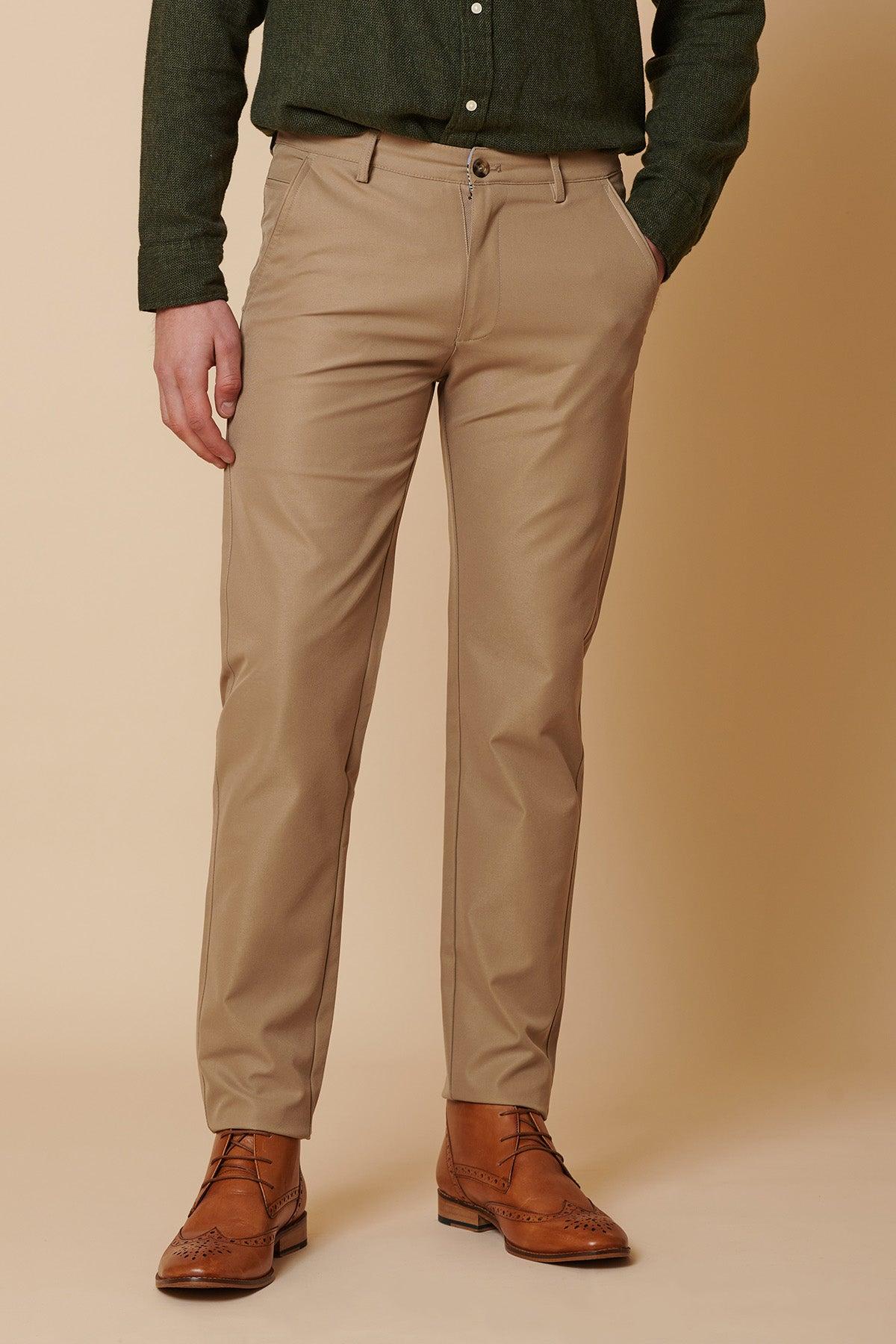 A person models the marcdarcy ENZO - Sand Chinos with hands in pockets, paired with a dark green shirt and brown leather shoes against a beige background. Their face is not visible in the image.