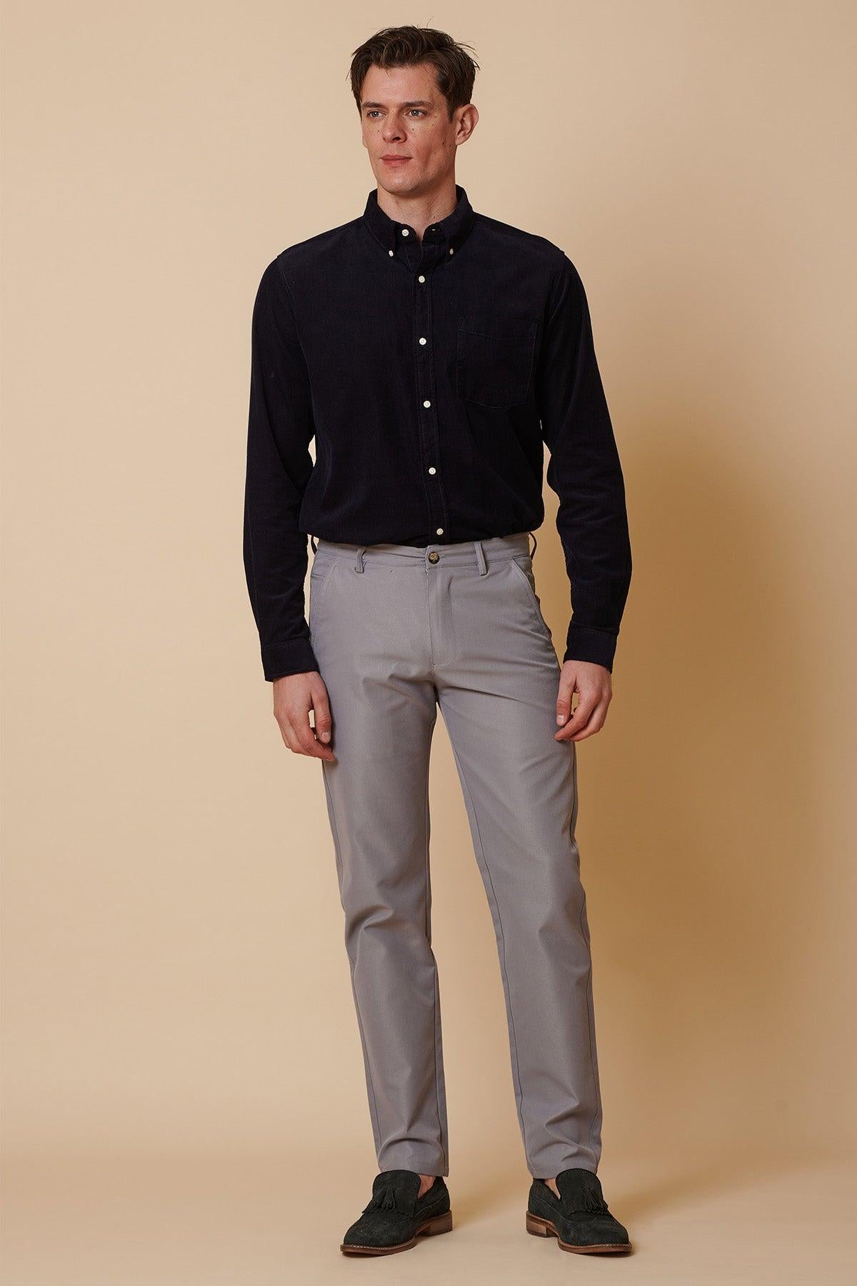 A man with short brown hair and a neutral expression stands against a beige background, wearing marcdarcy ENZO Mid Grey Chinos, a dark navy button-down shirt, and dark loafers.