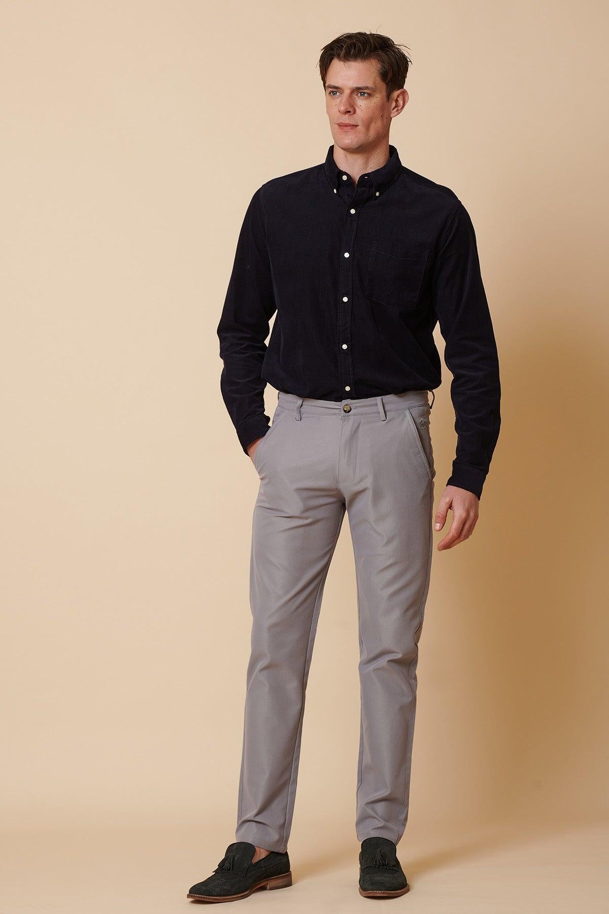 A man stands against a beige background, wearing marcdarcy ENZO Mid Grey Chinos, a black button-up shirt, and dark loafers. He looks slightly left with his left hand at his side and his right hand in his pocket.