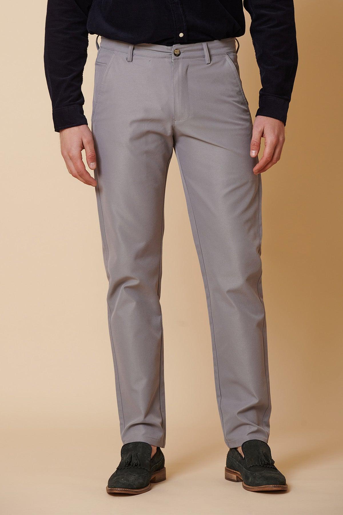 A person stands against a beige background wearing marcdarcy's ENZO Mid Grey Chinos, paired with a dark long-sleeve shirt and tassel loafers. Their head is not visible in the image.
