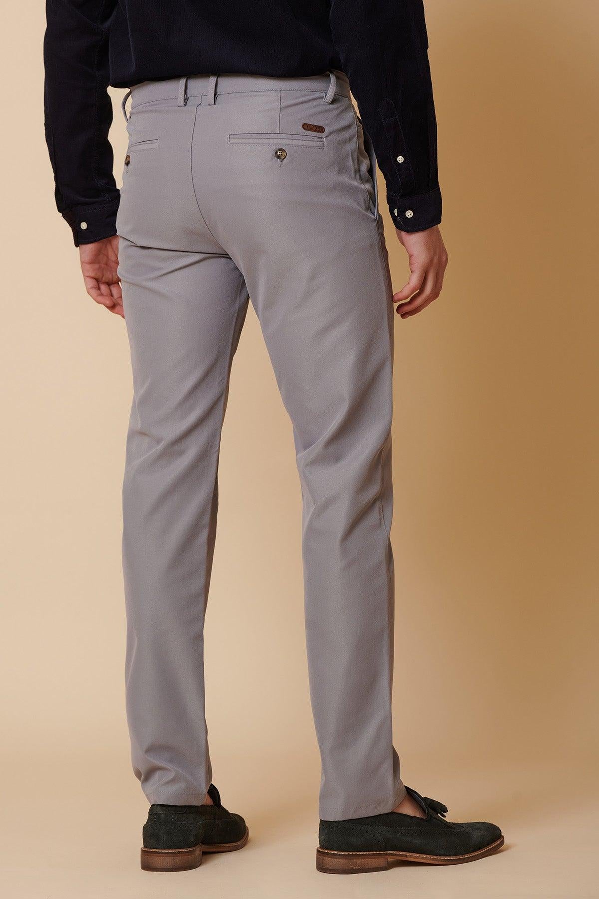 A man stands facing away against a beige background, wearing marcdarcy’s ENZO Mid Grey Chinos, a black shirt with buttoned cuffs, and dark green loafers.