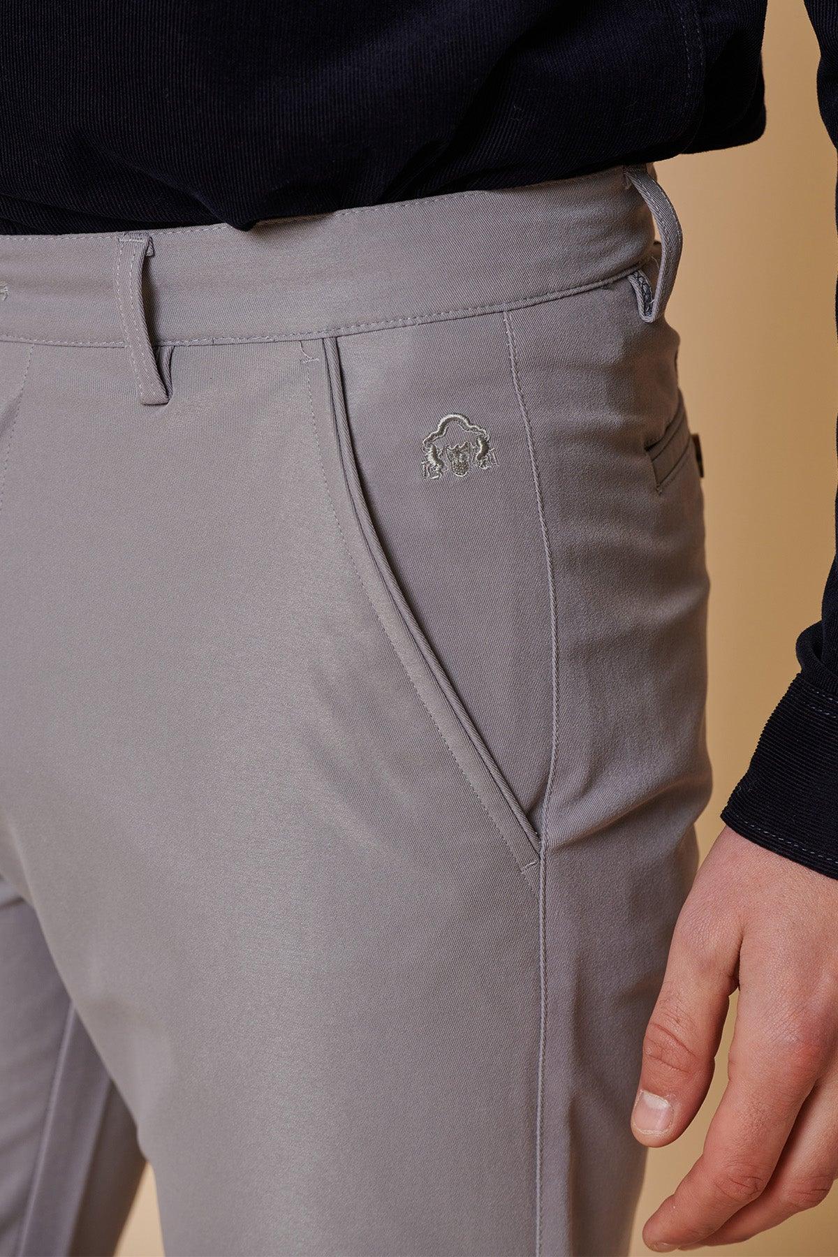 Close-up of marcdarcy ENZO mid grey chinos featuring slanted front pocket and small embroidered logo, styled with a navy top. Set against a beige background.