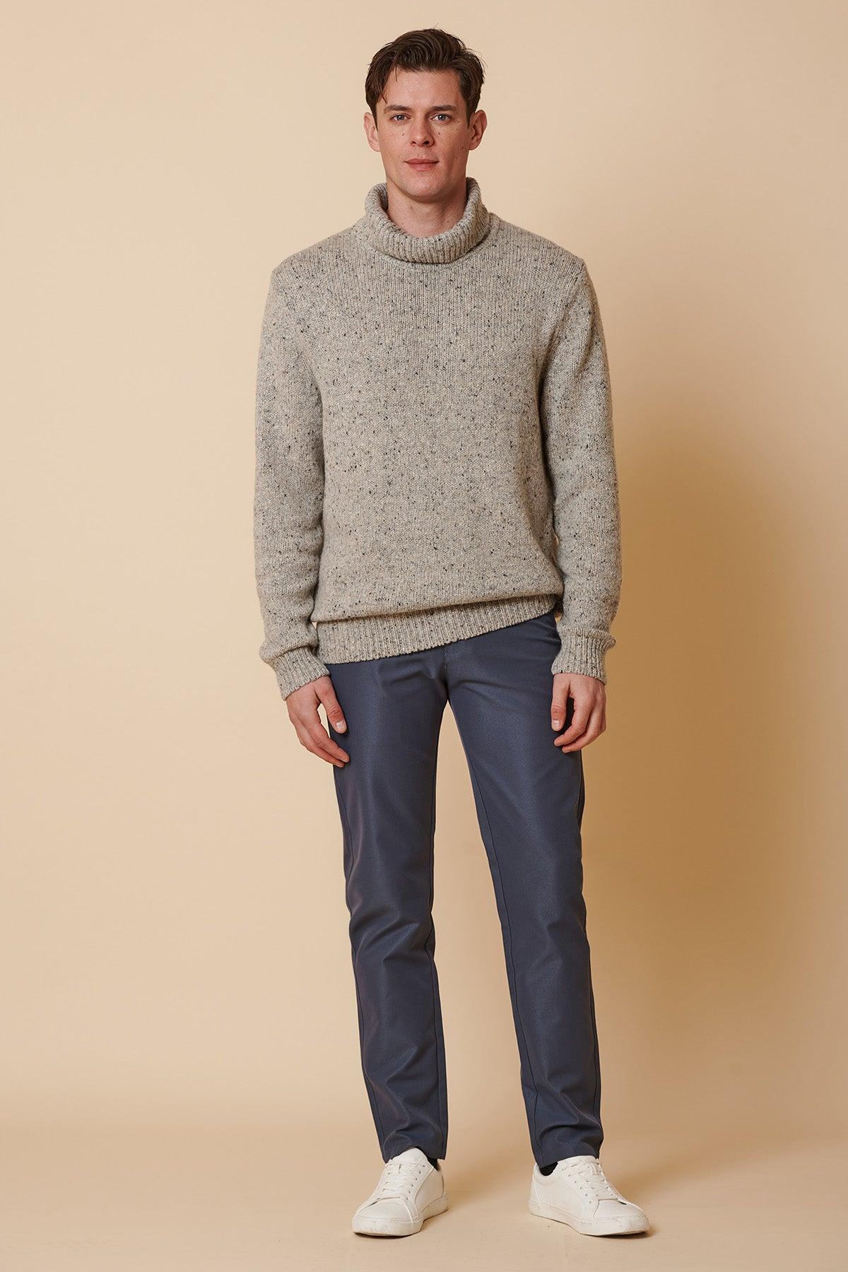 A man stands against a beige background wearing marcdarcy's ENZO Grey Blue Chinos, a gray turtleneck sweater, and white sneakers, facing forward with his arms by his sides.