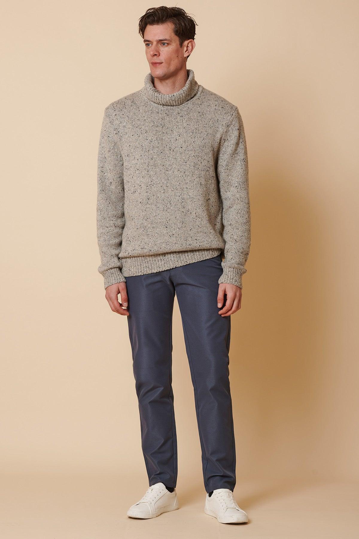 A man stands against a beige background wearing marcdarcy ENZO Grey Blue Chinos, a light gray turtleneck sweater, and white sneakers. He looks slightly to the side with a neutral expression.