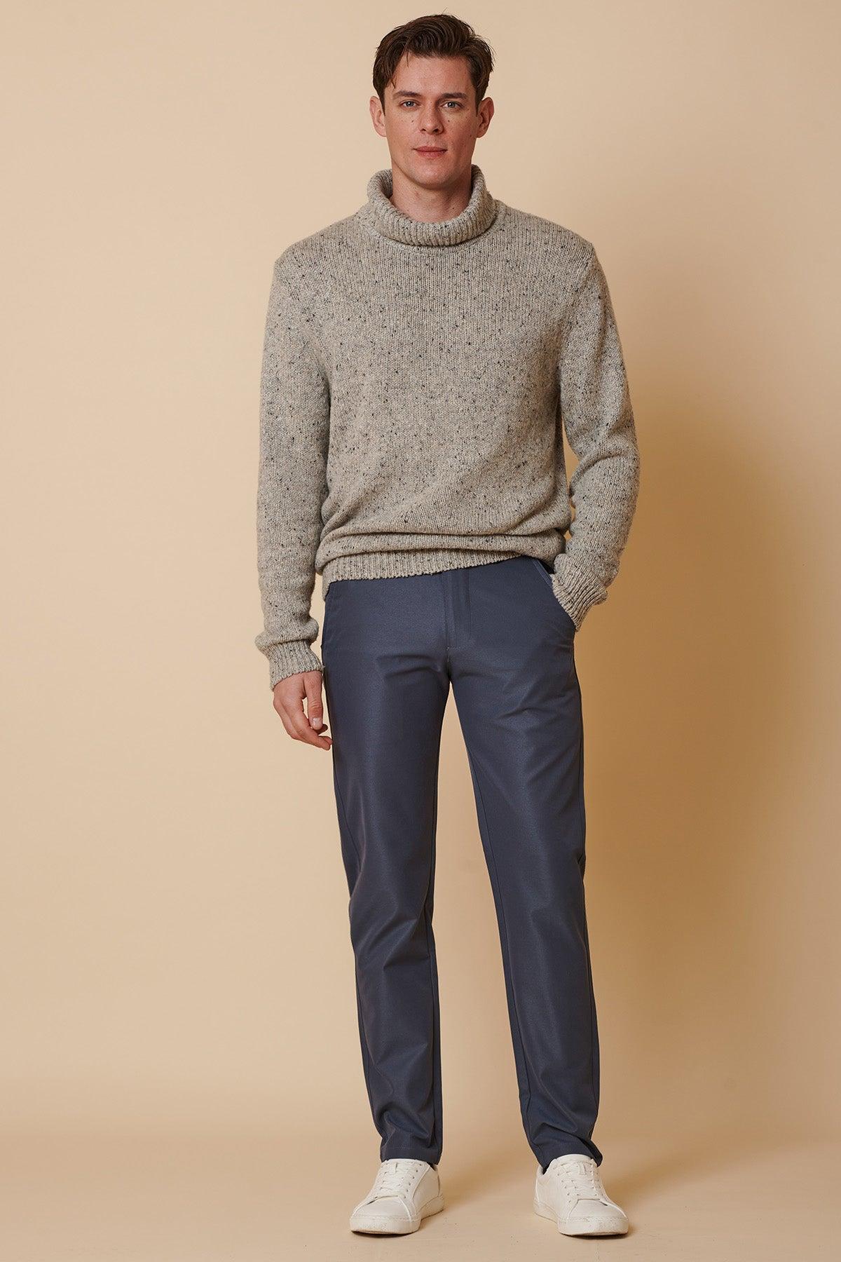 A man with short brown hair and a neutral expression stands against a beige background wearing marcdarcy's ENZO - Grey Blue Chinos, a gray turtleneck sweater, and white sneakers, with his hands relaxed at his sides.