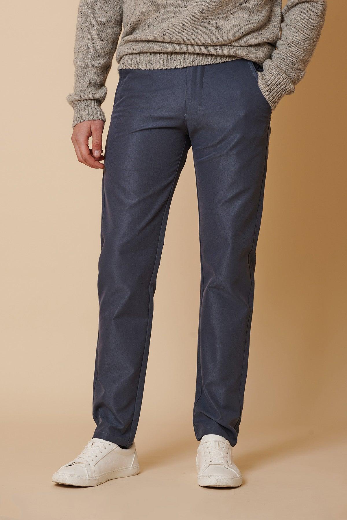 Wearing marcdarcy's ENZO Grey Blue Chinos, a person pairs them with a grey knit sweater and white sneakers, standing against a beige background with one hand in their pocket. The image is cropped from the shoulders down.
