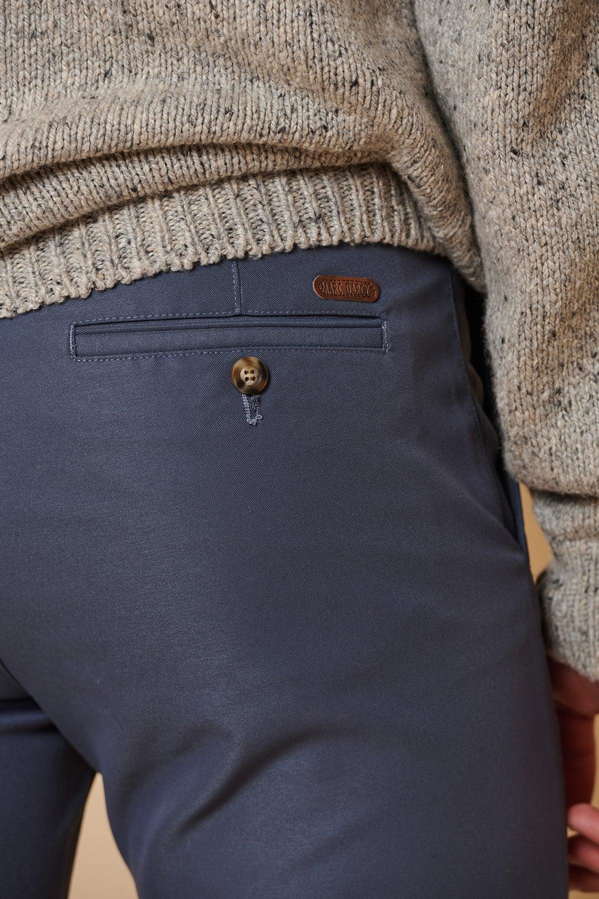 Close-up of a person wearing marcdarcy ENZO - Grey Blue Chinos featuring a buttoned back pocket with a small brown label, paired with a gray knit sweater. The person's arm is partly visible.