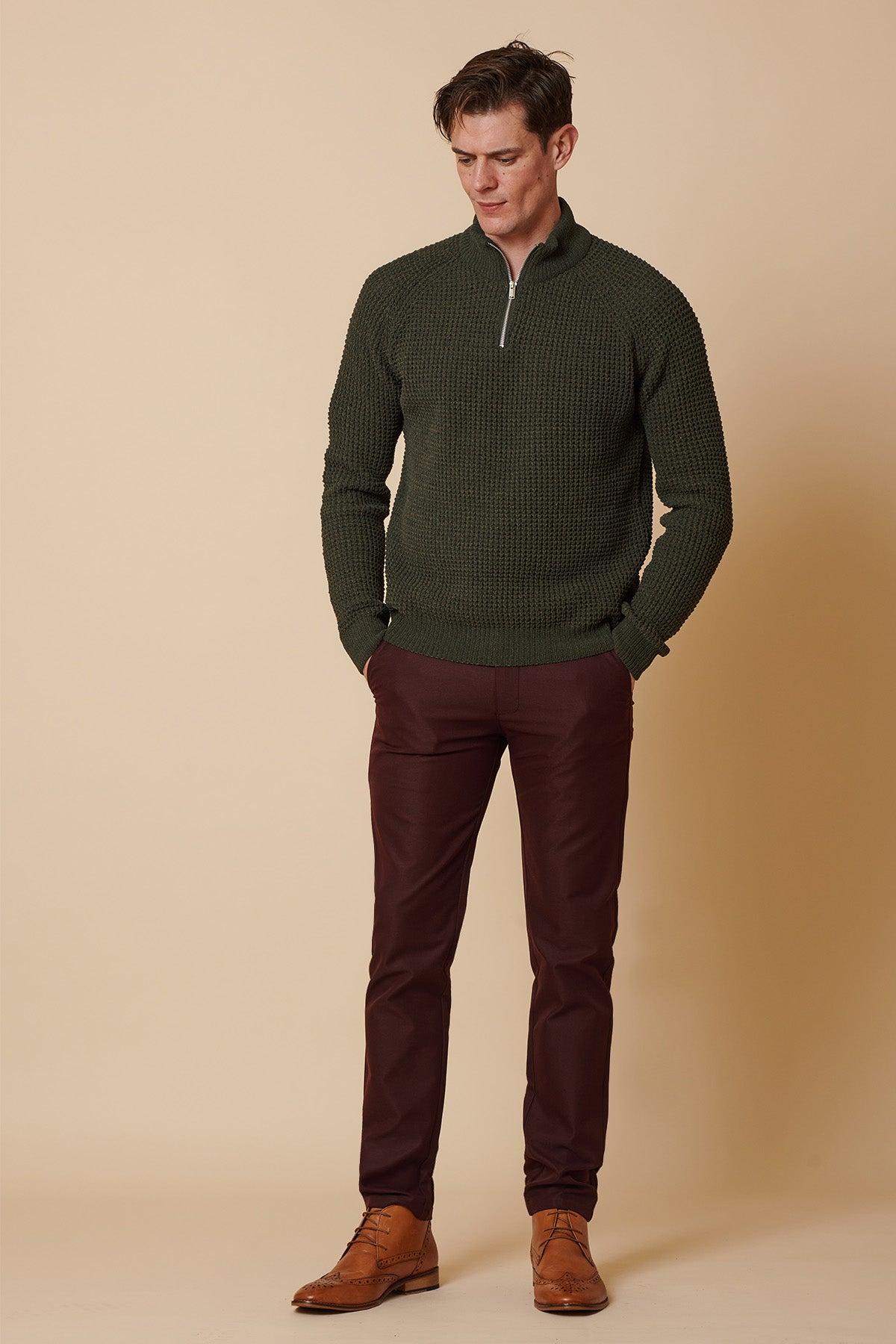 A man stands against a beige background, wearing marcdarcy's ENZO - Wine Chinos with a textured dark green quarter-zip sweater and brown dress shoes. His hands are in his pockets as he looks slightly downward.