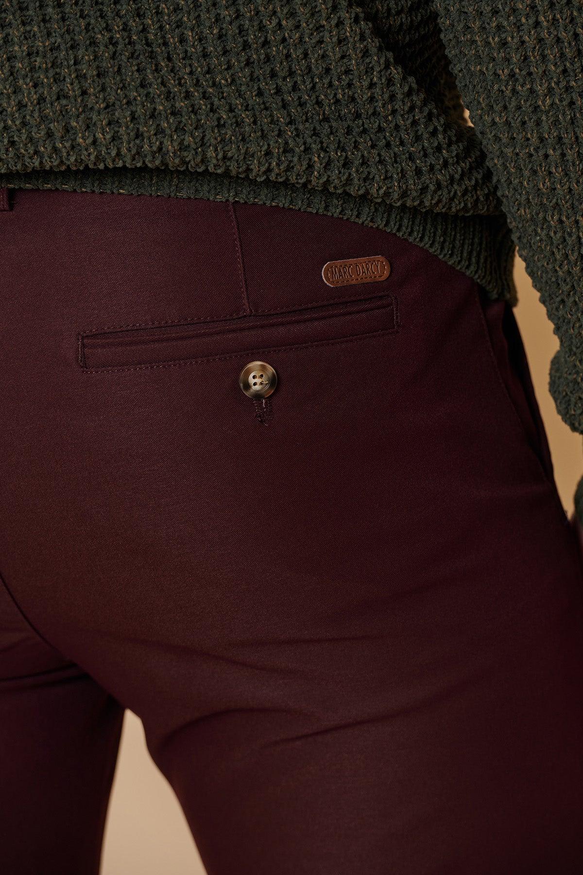 Close-up of a person wearing marcdarcy ENZO - Wine Chinos with a buttoned back pocket and a dark green knit sweater. A small oval leather patch with embossed text is visible above the pocket.