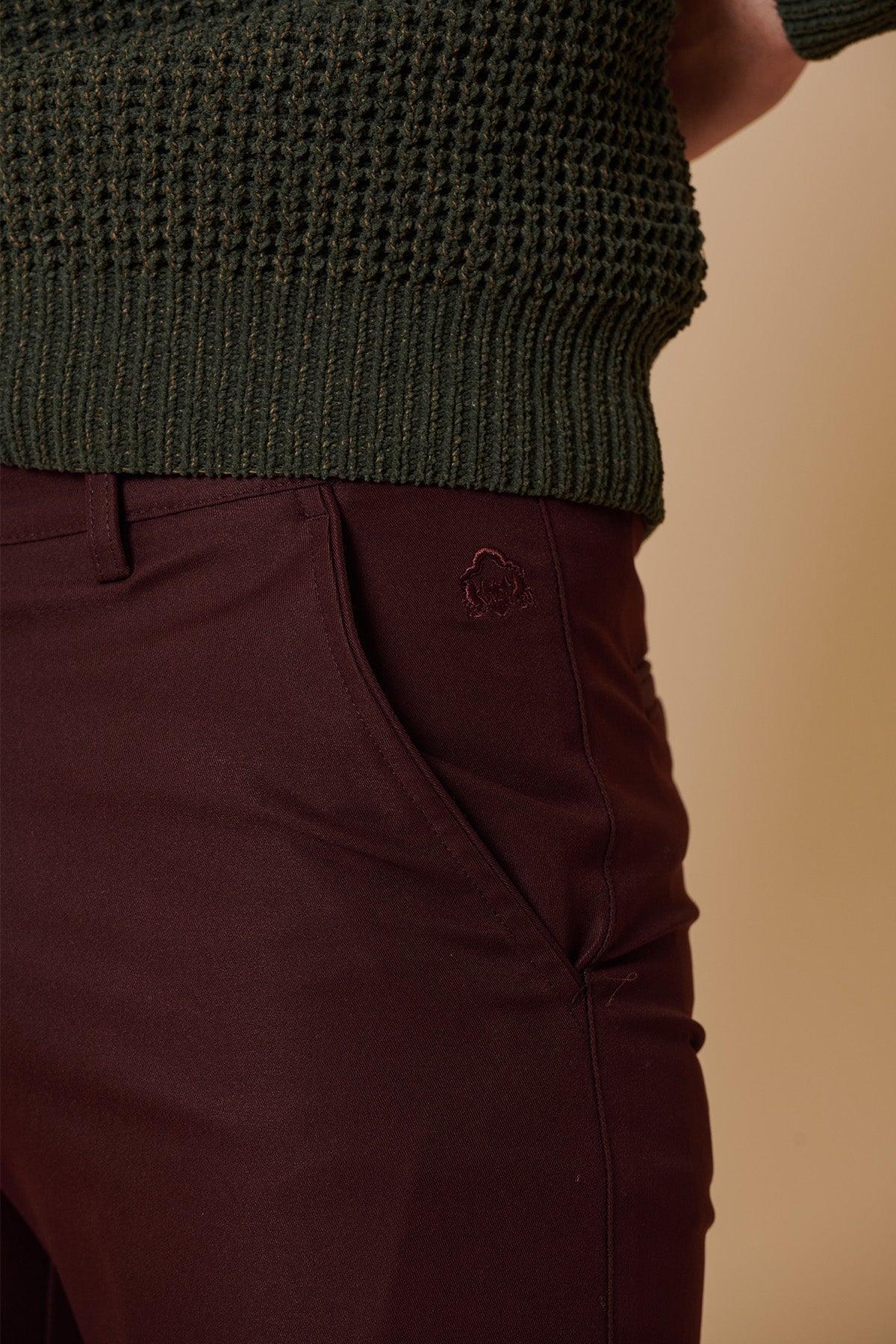 A person wearing a dark green textured sweater and maroon marcdarcy ENZO - Wine Chinos, featuring an embroidered emblem near the pocket, stands against a beige background.