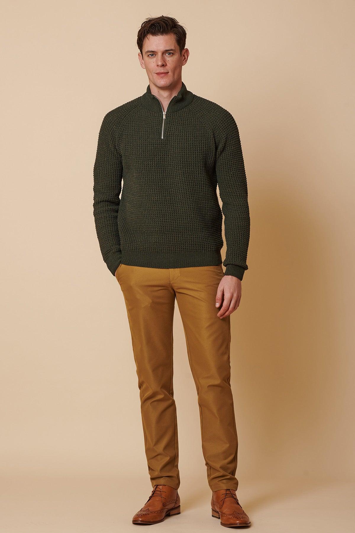 A man in a dark green waffle-knit quarter-zip sweater and marcdarcy ENZO Tan Chinos poses with his hands in his pockets against a light beige background.